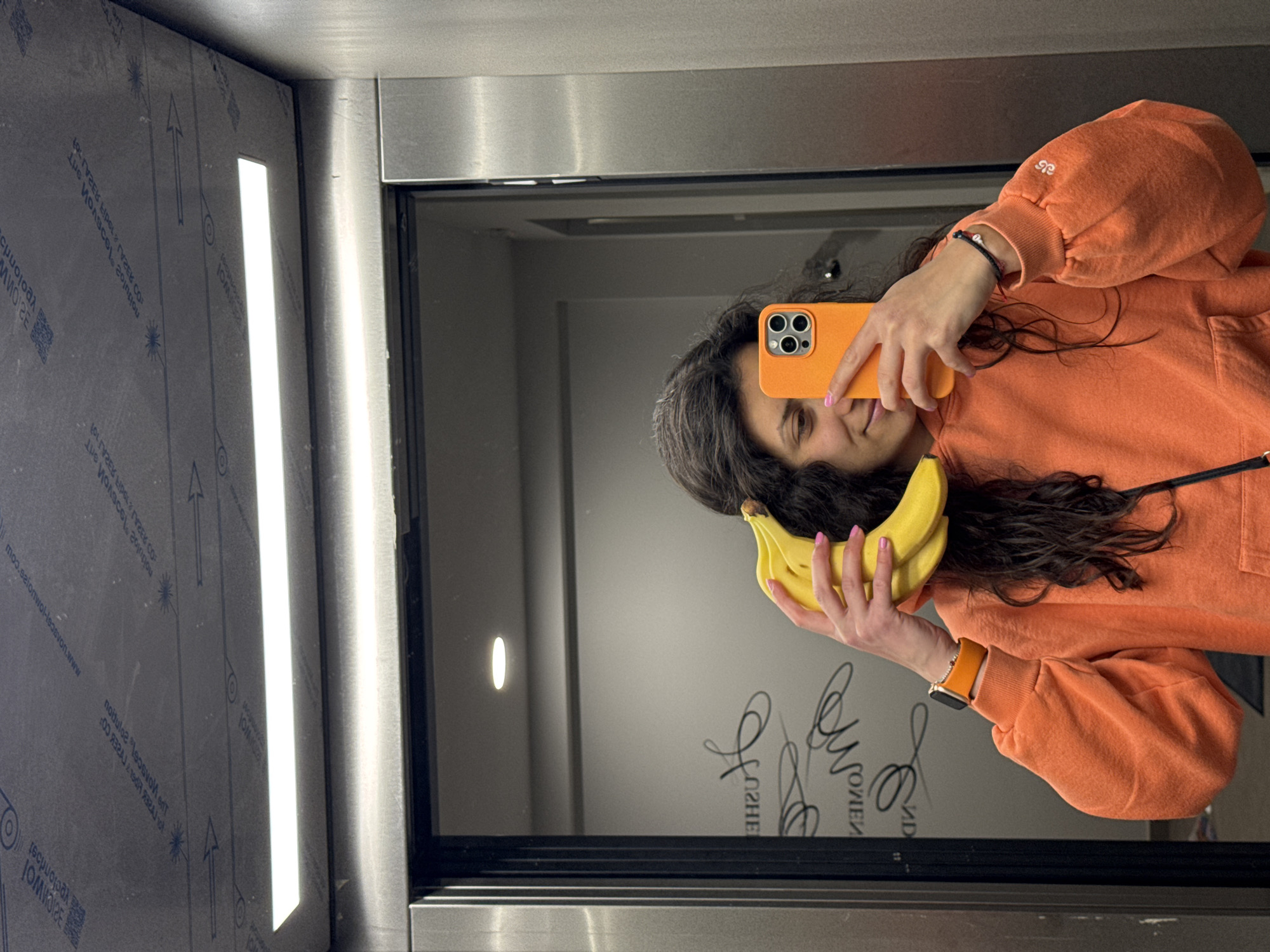 The image is a mirrored selfie of a woman inside an elevator. She is holding a bunch of bananas in one hand and an orange-cased smartphone in the other, presumably taking the photo. The woman is wearing an orange sweatshirt and has dark, curly hair. The elevator interior has a vertical light fixture on one wall and reflective panels. In the background, visible through the mirror, is an interior space with a light fixture on the wall and decorative writing. There is no clear indication of this scene’s location within Sofia, Bulgaria.