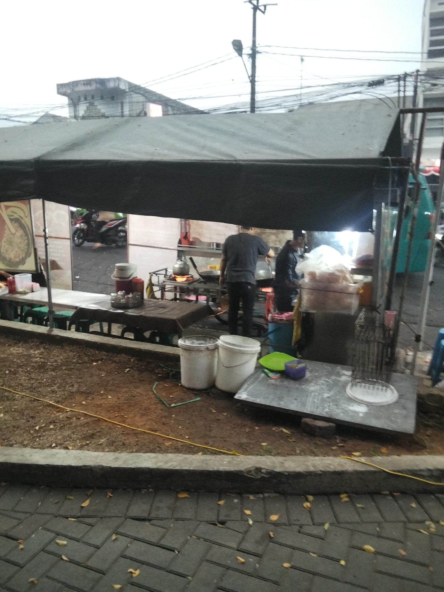 The image depicts an outdoor food stall in Cisaat, Indonesia. The stall is covered by a large, dark tarp. Two individuals are visible within the stall; one, wearing a dark t-shirt and black pants, is standing and appears to be cooking, with a pot on a stove in front of him. The other individual, wearing a dark jacket and dark pants, is facing away from the camera and also seems to be engaged in food preparation.
In the foreground, there is a paved area with dark, rectangular bricks. A yellow cord runs along the edge of this paved area. Scattered on the ground are dry, fallen leaves. Beyond the paving, there is a dirt area where several white buckets and some green and purple plastic items are placed on a flat surface.
The stall itself has tables covered with brown cloths, some of which are holding various containers and a kettle on a stove. Behind the stall, a motorcycle is visible, suggesting it is on a street or in a public area. The background shows a cloudy sky and buildings, including what appears to be a multi-story structure. The overall scene suggests a typical street-side food vendor operating in an urban environment.
