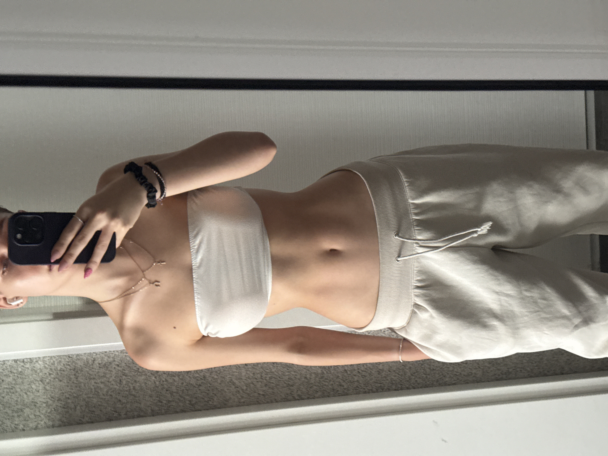 The image shows a person taking a selfie in a mirror. The person is wearing a white bandeau top and light-colored sweatpants. They have bracelets on their left wrist and a necklace with a cross pendant. Their fingernails are painted pink. The background appears to be a room with carpeted flooring and a white wall. The lighting suggests it is daytime. There is no visible text in the image. The setting is likely a home in Sofia, Bulgaria.