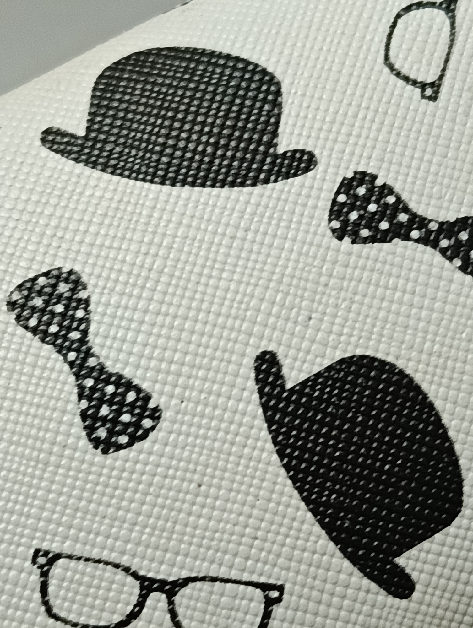 The image is a close-up, angled shot of a white textured fabric with a black pattern. The pattern consists of several stylized illustrations of men's accessories. On the upper left, there is a bowler hat. To the right of the hat, there are two bow ties with white polka dots. Below the bowler hat and to the left, there is another bow tie with white polka dots. In the lower right quadrant of the image, there is a pair of glasses with thick frames. The fabric's texture is a repeating grid of small raised squares, giving it a slightly bumpy appearance. The lighting is even, and the focus is sharp on the pattern. There are no people or actions depicted. The image is a still life of a fabric print and does not contain any information about its location in Biliaivka, Ukraine.