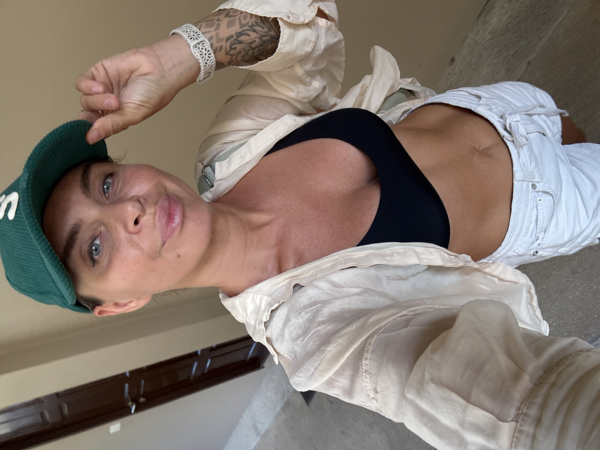 A woman, wearing a green baseball cap, a light-colored unbuttoned shirt over a black bikini top, and white shorts, poses for a selfie indoors. She is touching the brim of her cap with her right hand. A white patterned bracelet is visible on her right wrist, and tattoos are visible on her forearm. The background shows a doorway and a tiled floor, suggesting an indoor hallway or corridor. The lighting appears to be natural daylight. The setting is the island of Ko Samui in Thailand.