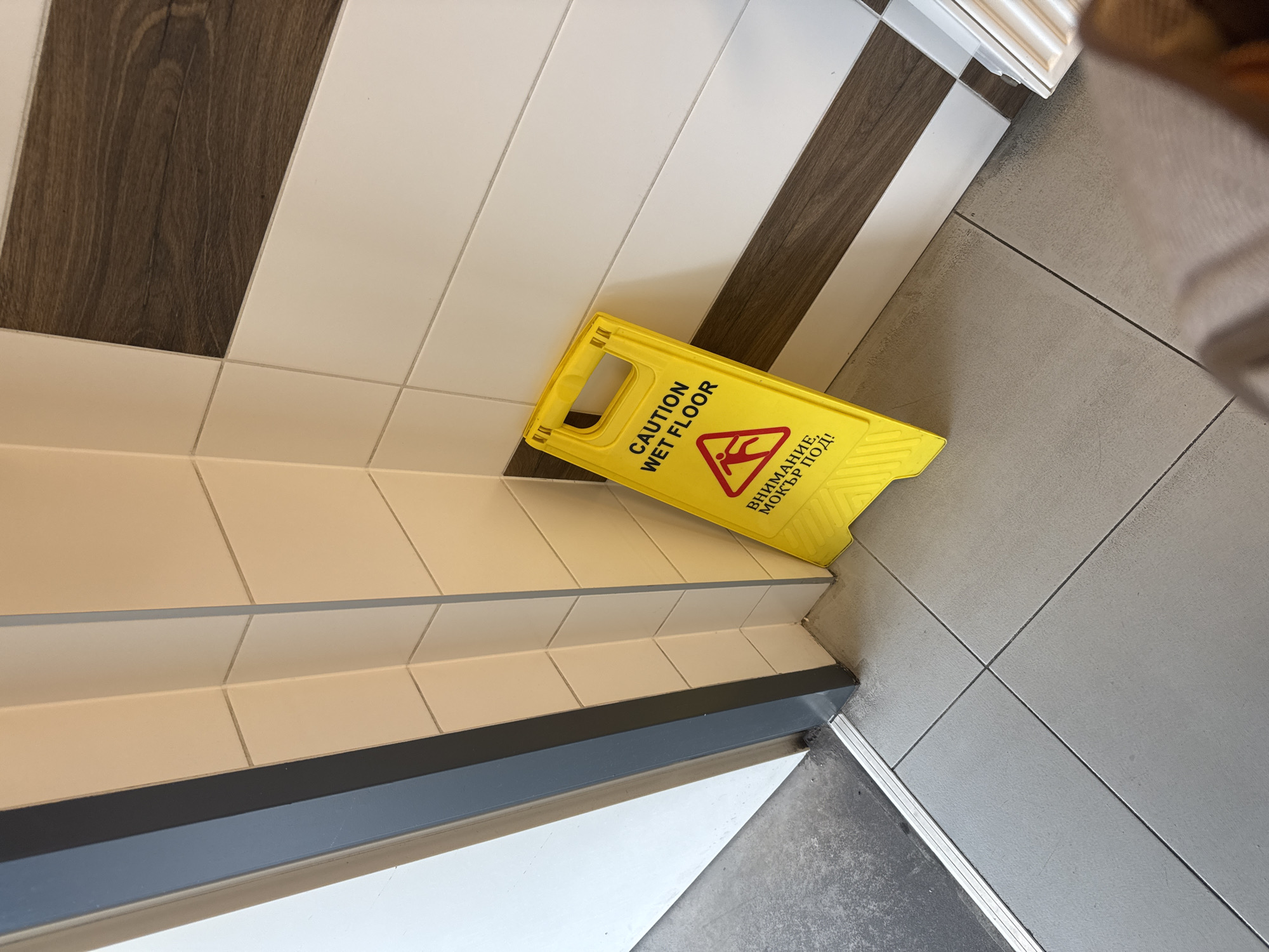 The image captures an indoor scene featuring a prominent bright yellow "CAUTION WET FLOOR" sign. The sign is triangular, displaying a red warning symbol of a person slipping, and includes text in English: "CAUTION WET FLOOR," as well as in Bulgarian Cyrillic: "ВНИМАНИЕ, МОКЪР ПОД!" which translates to "ATTENTION, WET FLOOR!"
The setting suggests a transition area within a building, likely in Plovdiv, Bulgaria, given the visible Bulgarian text. The flooring in the scene is multi-textured and patterned. In the upper portion, there's a distinct pattern of alternating white and dark brown, wood-grain-textured planks. This area transitions into a series of steps or a raised platform covered in light-colored, possibly cream-toned, square tiles. A dark grey or black protective trim runs along the edge of these steps. Further down and to the right, the flooring shifts again to a grey tiled surface with visible grout lines, which meets another darker surface via a metallic transition strip.
The presence of the sign indicates that the floor in the immediate vicinity is wet, likely due to cleaning or a spill, and advises caution to prevent slips and falls. No people are visible in the image, but the sign implies recent human activity related to maintenance or an incident. The lighting suggests an interior environment.