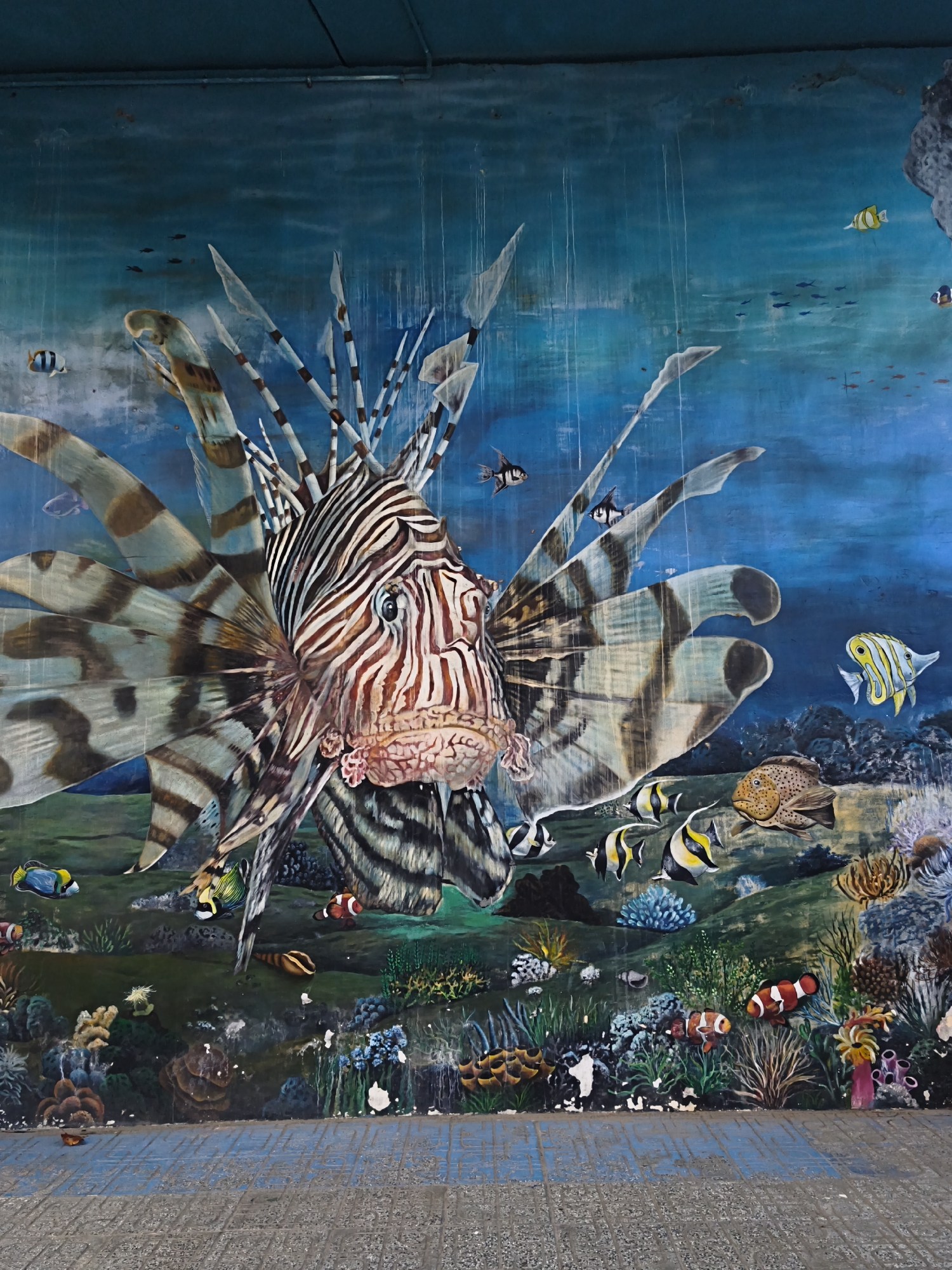 This is a vibrant mural depicting an underwater scene with a large lionfish as the central subject. The lionfish, rendered with detailed stripes and flowing fins, dominates the foreground. It is surrounded by a variety of smaller, colorful fish, including angelfish, clownfish, and damselfish, swimming among coral reefs, anemones, and sea grass. The background suggests the open ocean with a deep blue, textured wash that appears to mimic water. Streaks of white paint create the illusion of falling water or light rays.
The scene is a lively and dynamic representation of marine life, likely intended to evoke a sense of wonder and the beauty of the ocean. There are no people visible, and the mural itself is the main object. The setting is clearly an underwater environment. There are no discernible time of day or weather cues, as it is a painted representation. Location cues are limited to the information provided about Bac Nha Trang Ward, Vietnam, suggesting this mural is part of the local scenery or public art in that area. The overall emotion conveyed is one of natural beauty and perhaps a touch of awe due to the dramatic portrayal of the lionfish. No text is visible in the image.