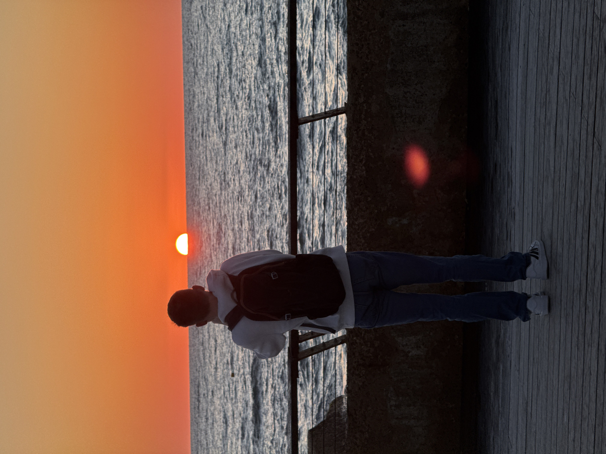 The image depicts a tranquil sunset scene over a body of water, possibly a sea or ocean. A single individual stands on a dark wooden deck or pier, viewed from behind, gazing towards the horizon. The person is wearing a light-colored hoodie, blue jeans, and white sneakers, and has a dark backpack on. A dark metal railing runs across the mid-ground, separating the deck from the water.
The sky is a vibrant gradient of orange and yellow, characteristic of sunset, with the bright, partially visible sun positioned near the horizon line. Below the colorful sky, the water appears dark and textured with gentle waves, reflecting some of the ambient light. The person and the railing are largely silhouetted against the bright backdrop of the setting sun. A red lens flare is noticeable in the darker areas of the image, particularly on the right side. The overall mood is one of peaceful observation, as the person watches the sun descend. There is no visible text in the image.