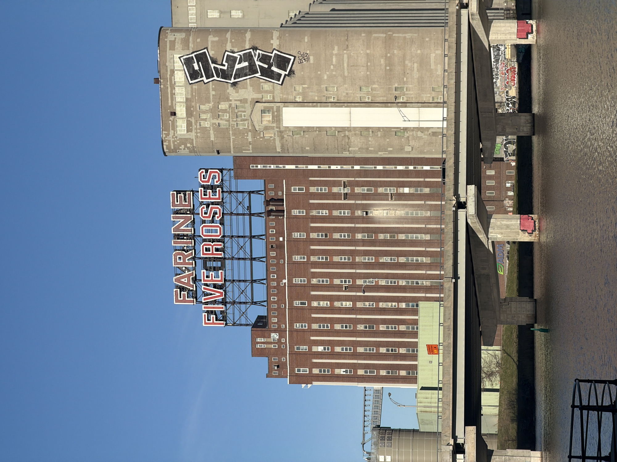 The image depicts a collection of industrial buildings and structures in Montreal, Canada, under a clear blue sky. Prominently featured is the Farine Five Roses sign, a large vintage neon sign atop a brick building, a well-known landmark in the city. Adjacent to the brick building is a tall concrete grain elevator covered in graffiti. A bridge structure is visible on the right side of the image, with concrete supports and graffiti underneath. The water of the Saint Lawrence River is visible in the foreground, reflecting the surrounding structures.