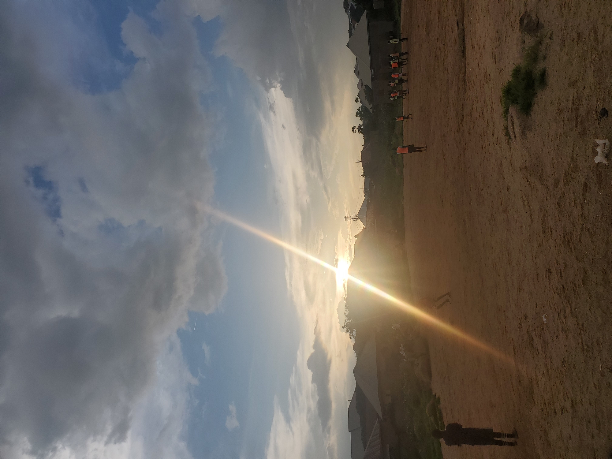 A wide shot captures a dirt field where a group of young people, some wearing orange vests, are engaged in a soccer game. The sun is low in the sky, casting a strong lens flare across the scene and illuminating the clouds above. In the background, a cluster of homes with peaked roofs is visible, suggesting a residential area. A lone figure stands with their back to the camera on the left side of the field. The overall atmosphere is that of an outdoor recreational activity in a rural or semi-rural setting.
