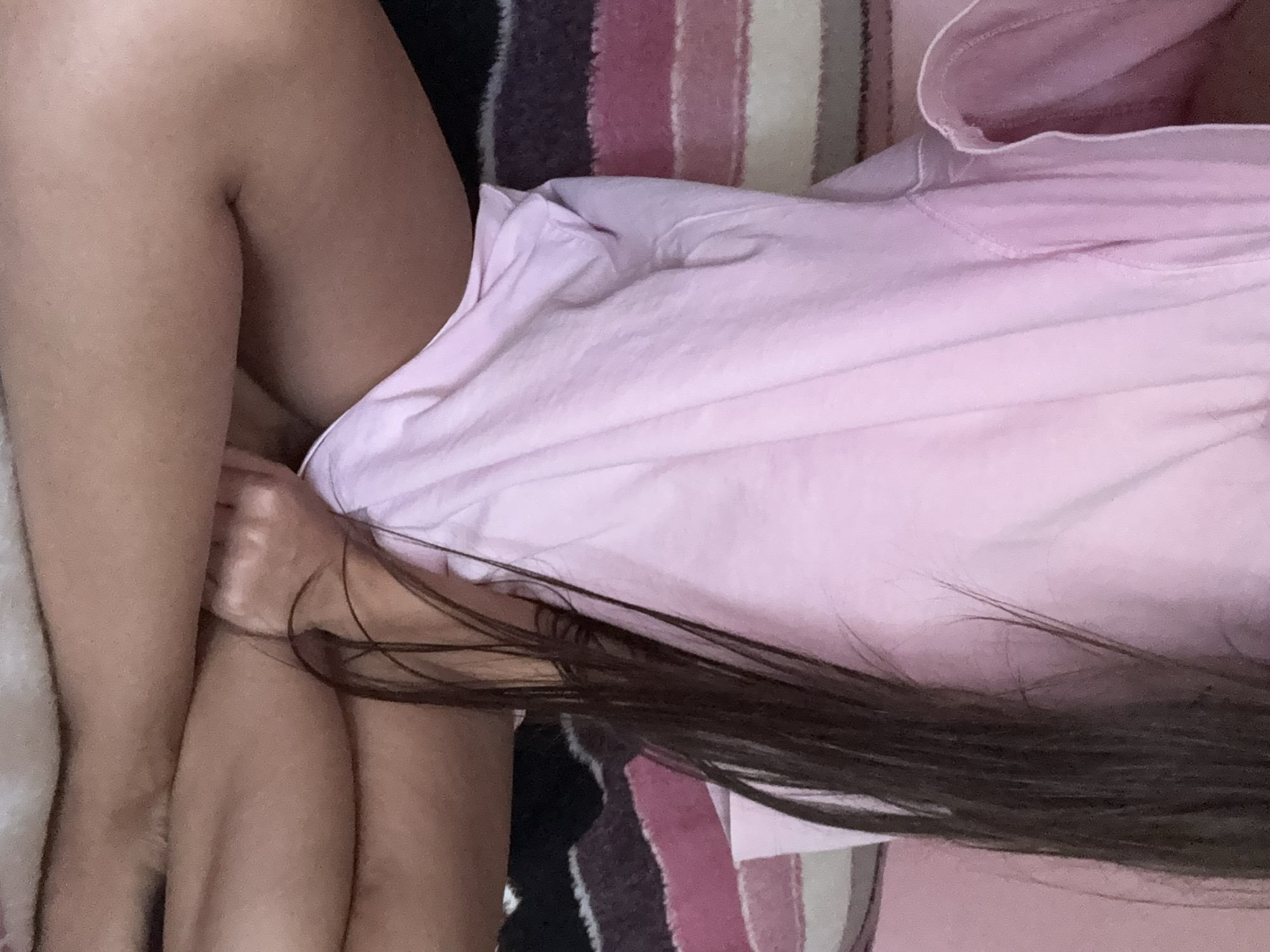 The image shows a close-up of a person's legs and torso, wearing a pink t-shirt and sitting cross-legged. The background is a striped blanket or cushion with stripes of pink, purple, and black. The person's long, dark hair is visible falling over their shoulder. There is no indication of the location in Basarabeasca, Moldova.