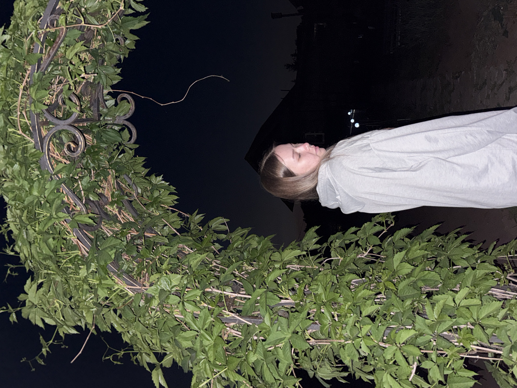 A nighttime scene shows a woman with long blonde hair wearing a white hoodie. She is positioned to the right of a wrought-iron archway that is heavily covered with green leafy vines. The background is dark, suggesting an outdoor location. The presence of building silhouettes and distant lights suggests an urban environment, potentially Zhigalki, Russia.