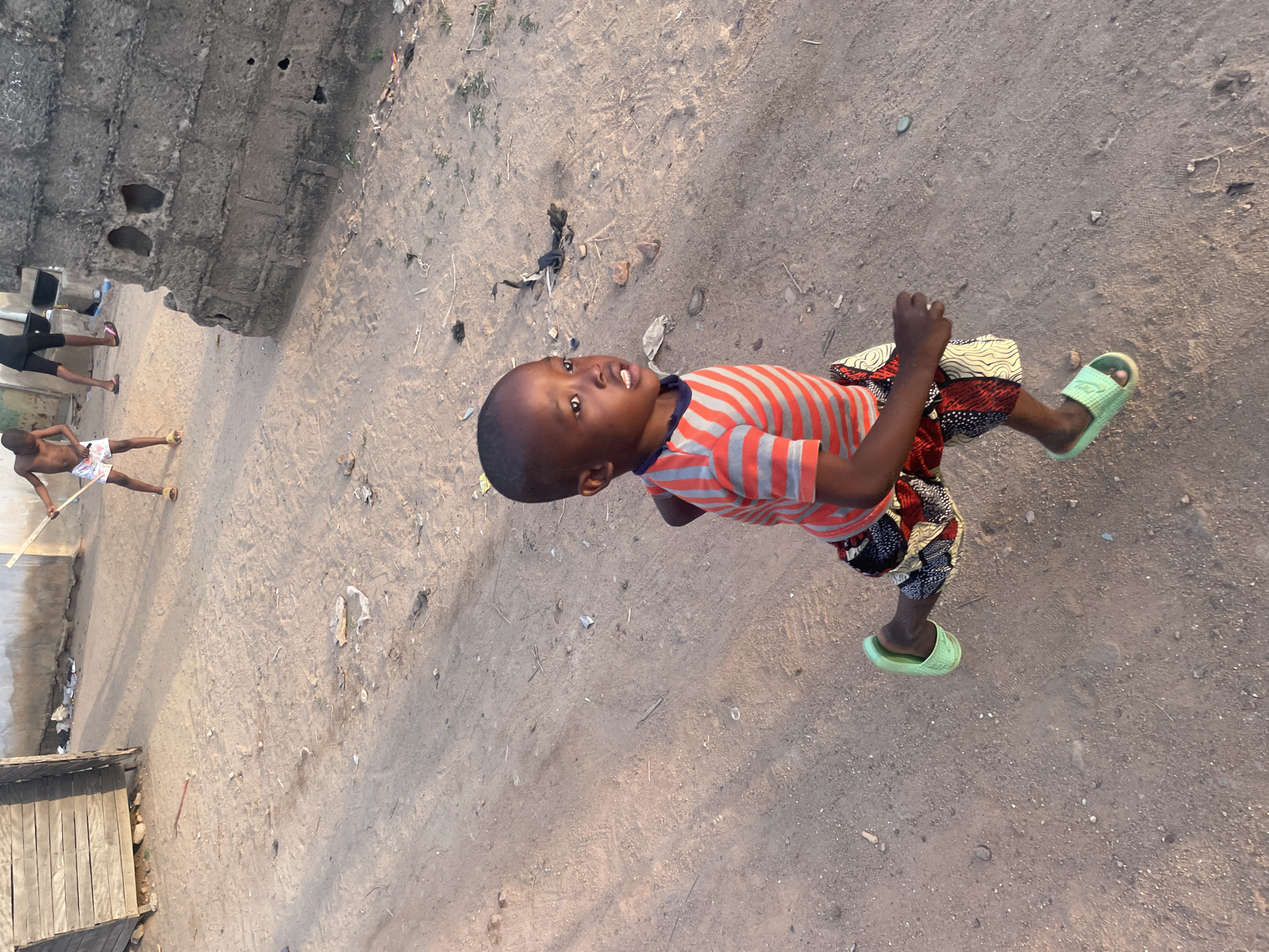 A low-angle, rotated medium shot depicts a child in Lokoja, Nigeria, standing on a dirt surface. The child wears a red and gray striped shirt, patterned shorts, and green sandals. They are looking up toward the camera. In the background, other figures are visible near a dilapidated concrete structure. The surrounding environment includes scattered debris and a rudimentary wooden structure.