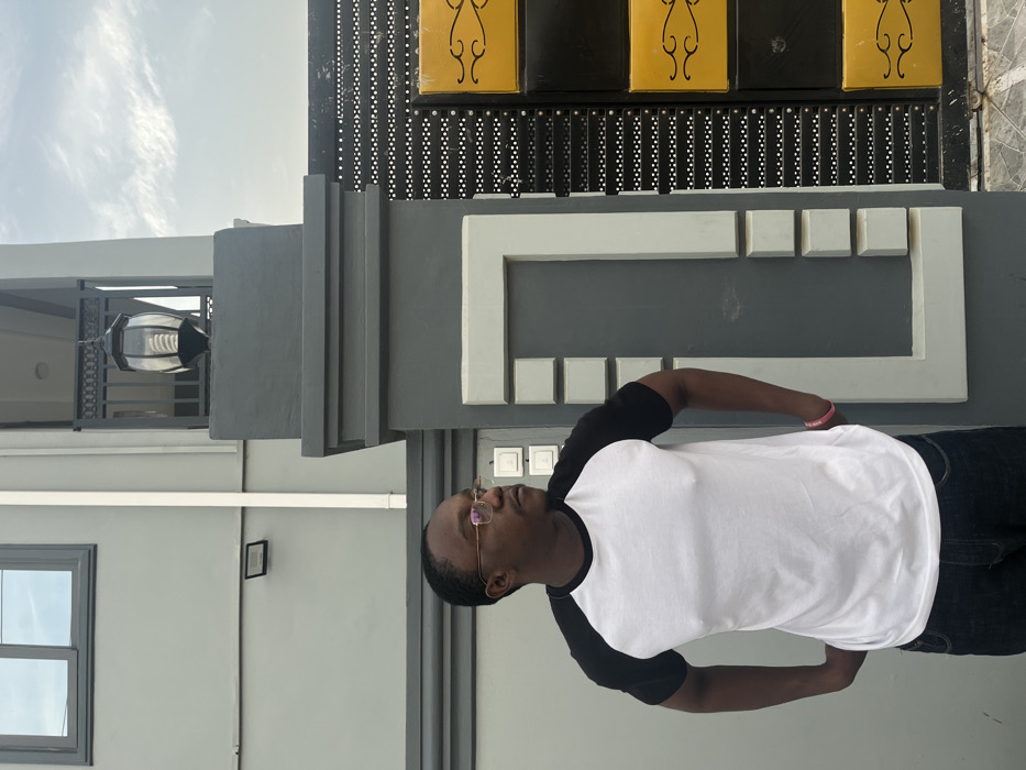 An outdoor, daytime shot shows a dark-skinned man standing in front of a modern building in Joma, Ghana. The man is wearing a white raglan t-shirt with black sleeves, dark jeans, and gold-rimmed glasses. He has a pink wristband on his right wrist and is looking upwards and slightly to the right with a neutral expression.
The background features a multi-layered architectural design. Directly behind the man is a dark gray wall adorned with a white, rectangular frame-like structure made of varying-sized blocks. Above this, a dark, perforated gate or fence panel is visible, topped by two black panels with yellow, ornate scrollwork decorations. To the man's left, a light gray wall features a dark-framed window, and a silver-colored outdoor lantern hangs from an overhang above a balcony. Another small, dark rectangular object is affixed to the light gray wall. The sky is bright with some scattered, wispy clouds, indicating clear weather. No visible text is present in the image.