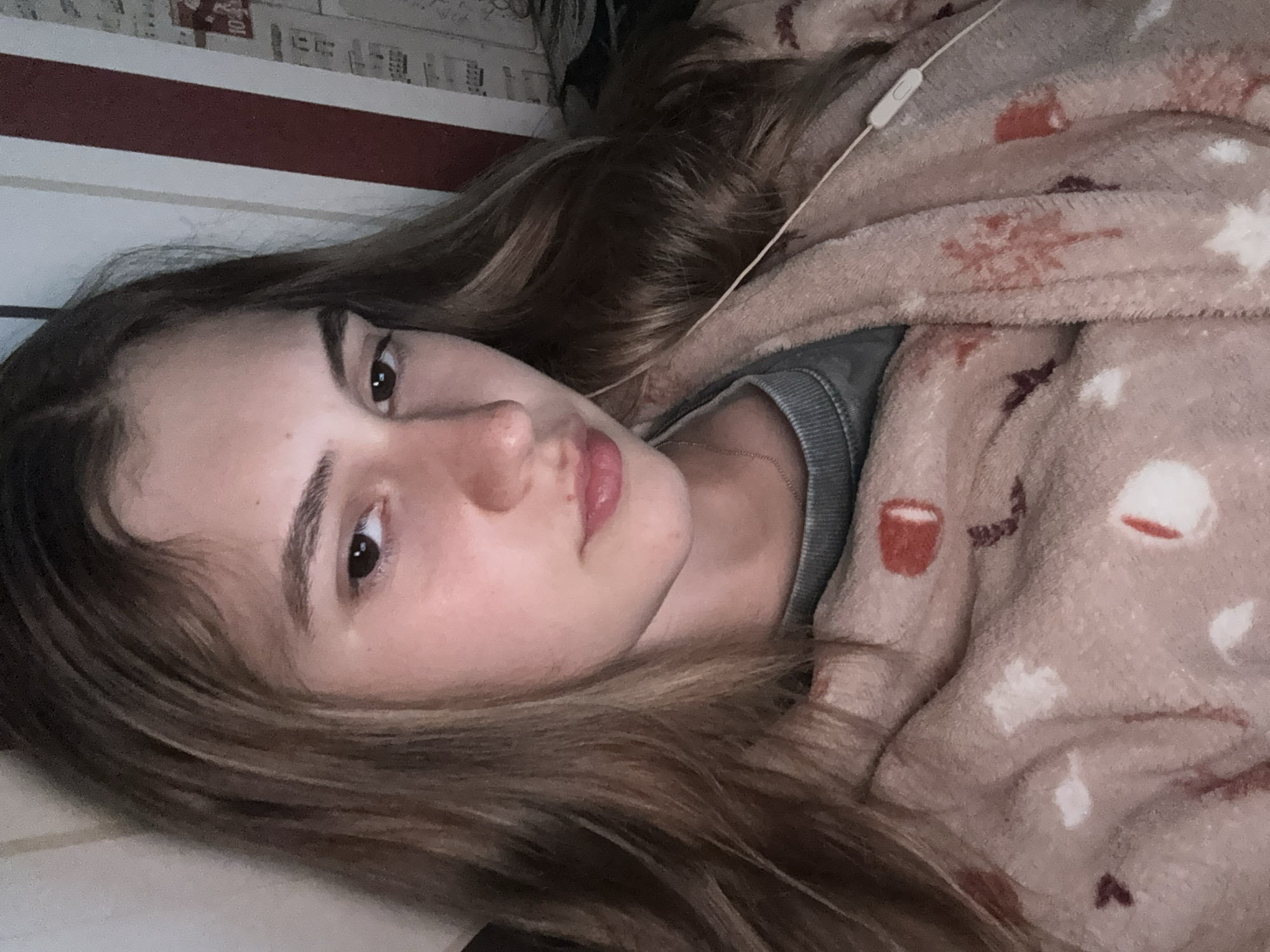 A young woman with long, light brown hair is lying down indoors, looking directly towards the camera. Her head is positioned to the left side of the frame (when viewed with the image rotated 90 degrees clockwise to an upright orientation). She has fair skin, dark eyes, and her lips show a hint of pink. Her expression is calm and neutral.
She is partially covered by a fluffy, light pink or beige blanket patterned with red and white shapes, some resembling coffee cups or abstract forms. Beneath the blanket, a grey top, possibly a sweater, is visible, along with a thin, gold-colored necklace chain around her neck. A white earbud cable, including an in-line control module, rests on the blanket near her head.
The background consists of a wall with a prominent horizontal red stripe. Above this stripe, the wall features a lighter color with subtle, indistinct patterns that appear to be small graphics or unreadable text. The setting suggests a casual, relaxed indoor environment, possibly a bedroom or living room. No specific location cues are visible within the image to confirm Panagyurishte, Bulgaria. The time of day is not discernible, but the relaxed posture implies a moment of rest or leisure. No clear, readable text is visible.