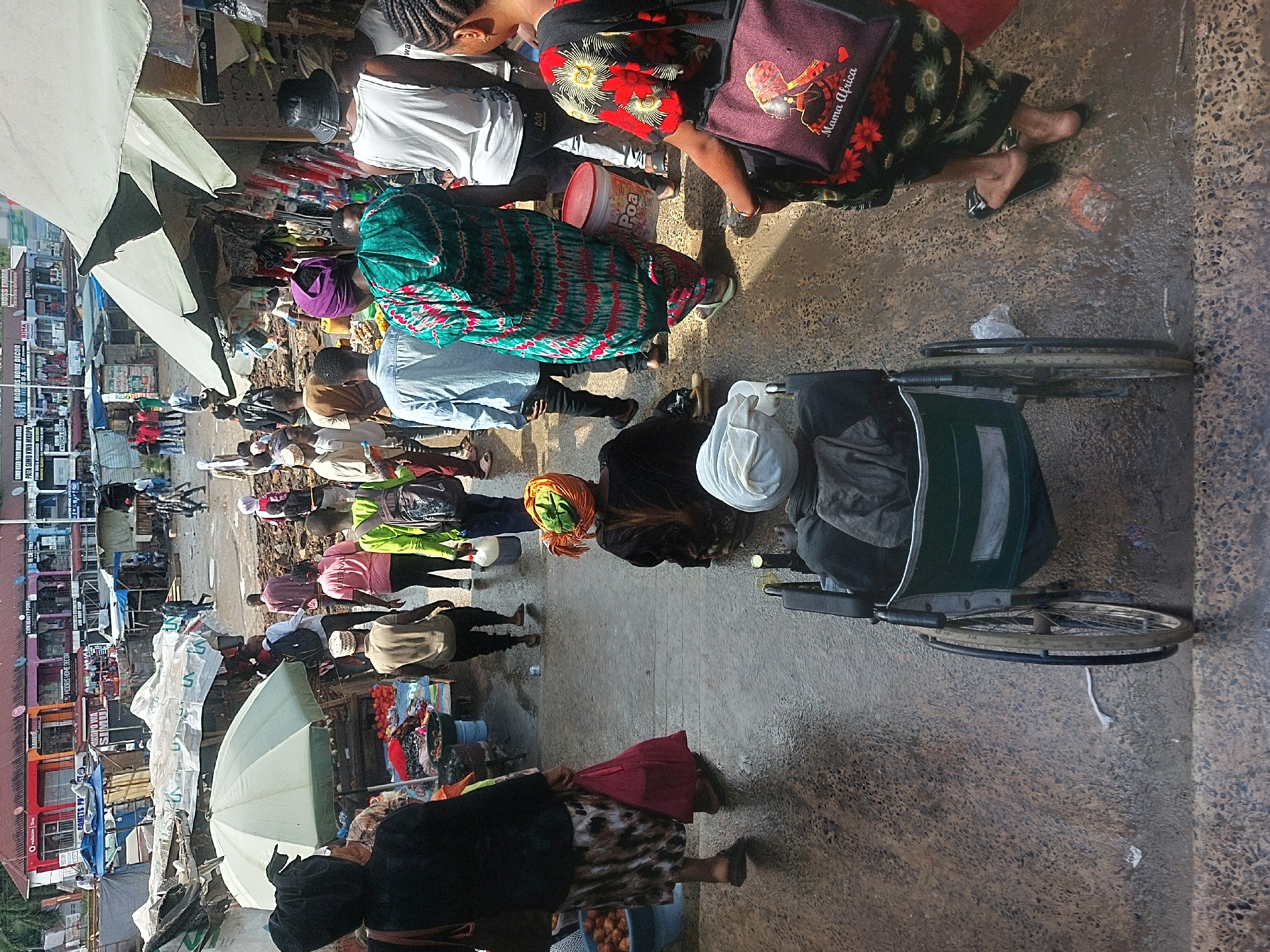 This media file captures a bustling outdoor market scene in Dar es-Salaam, Tanzania, filled with numerous people, market stalls, and urban architecture. The overall impression is one of a vibrant commercial hub.
The scene is densely populated with individuals of various ages, predominantly adults, engaged in walking, shopping, and selling. Many people are dressed in colorful, patterned garments characteristic of African attire, such as a prominent figure in a green and red patterned dress, and another with an orange headscarf. Several white and light green umbrellas are deployed, likely providing shade for vendors and their goods.
In the foreground and midground, there are multiple groups of people gathered around market displays, though the specific goods for sale are not clearly discernible beyond general market items. On the right side of the frame, there is a distinct tricycle or wheelchair with a large seat, positioned on the concrete ground. The ground itself is paved and shows signs of wear and possibly moisture.
The background features multi-story buildings housing various businesses, indicated by visible signage. Text cues throughout the image reinforce the urban market setting. Visible text includes "MODRIS HOME DECOR," "SIMU USED FROM DUBAI & KOREA MODIIS HOME DECOR," "PATA SIMU KWA," "KUTOKA DUBAI," "BOSS EMMA," "BIMA," and "BARBER ABERSHOP." A red bucket in the midground has "Poa" written on it, and a purple bag displays "Mama Africa."
The lighting suggests it is daytime, possibly late morning or early afternoon, with diffused light casting soft shadows. The atmosphere is active and dynamic, reflecting the daily commerce of a busy street market.