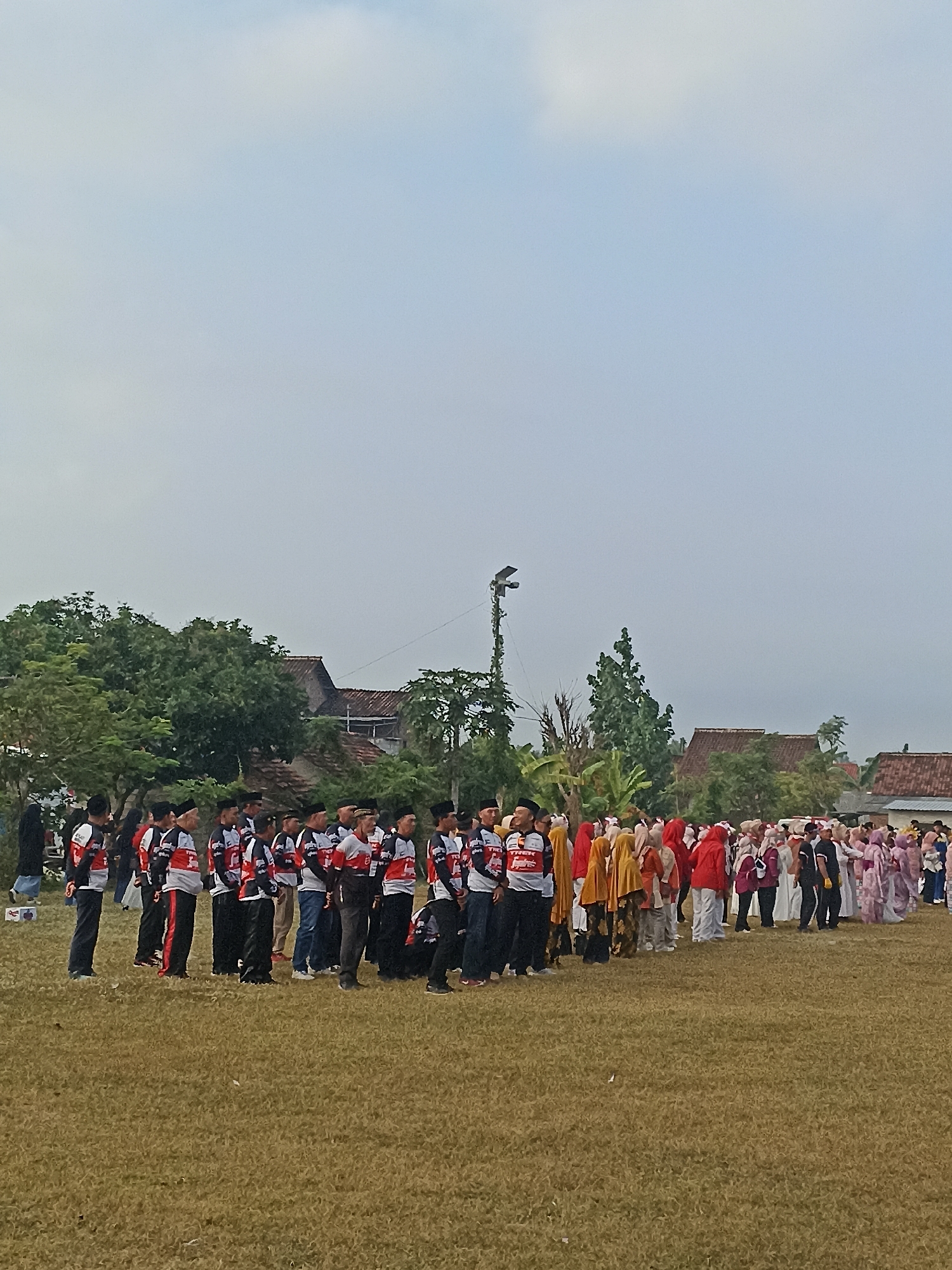 An outdoor, low-angle, full shot captures a gathering of people on a grassy field. The sky above is overcast and pale blue. In the background, a line of trees separates the field from a cluster of buildings with reddish-brown tile roofs, suggesting a rural or suburban setting. A tall, slender pole with a light fixture stands to the left of the frame.
The foreground and midground are occupied by two distinct groups of people standing in rows. On the left, a group of men is dressed in matching black and red sportswear with white accents, and many are wearing black skullcaps. To their right, a larger group, predominantly women and girls, are dressed in colorful attire, including yellow and red headscarves and flowing garments. The people appear to be facing forward, and some are looking towards the camera. The overall scene suggests an organized event or gathering, possibly a ceremony or festival, taking place in the city of Banyuwangi, Indonesia, given the style of dress and architecture.