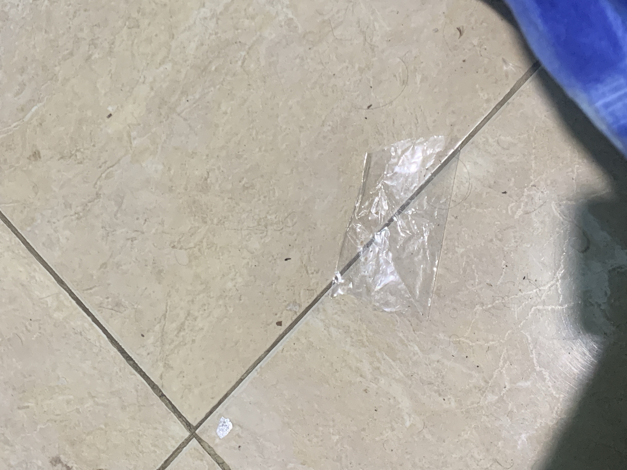 This close-up, high-angle shot captures a section of a light-colored tiled floor, likely beige or cream, in an indoor setting in Santa Ana, Venezuela. The tiles feature a subtle, irregular pattern resembling natural stone, and are delineated by dark grout lines that form a grid across the image.
The primary subject on the floor is a crumpled, clear plastic wrapper or small bag, positioned towards the center-right of the frame. Its transparency allows the underlying tile pattern to be partially visible. Scattered across the tiles and nestled within the grout lines are various small pieces of debris, including dust particles, tiny dark specks, and several strands of hair, some appearing curled. One specific dark, rectangular speck is visible, hinting at a small insect or a distinct piece of debris. Additionally, a small, irregular white mark, possibly paint or a piece of paper, can be seen near a grout line in the lower-left corner.
A blurry, dark blue object, likely made of fabric or plastic, is partially visible in the top right corner and along the right edge of the frame, suggesting it is a nearby item. The scene is static, portraying the floor and its scattered contents without any activity taking place. The lighting appears to be ambient indoor light. There is no visible text in the image.