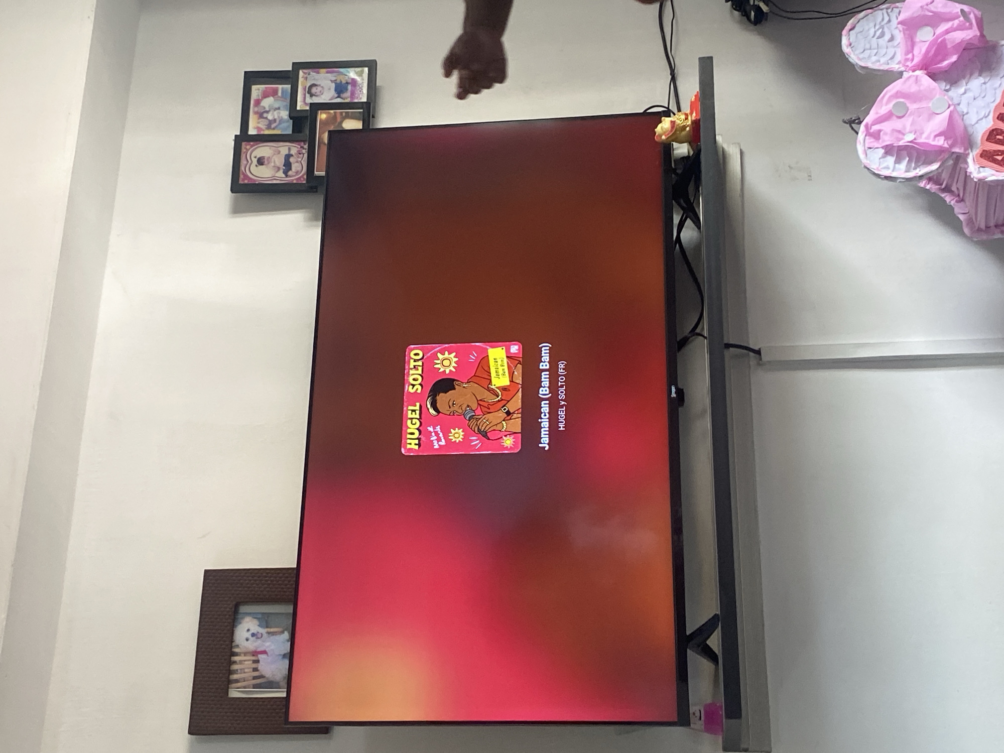 A wall-mounted television displays music by Hugel Solto: Jamaican (Bam Bam), next to various framed photographs. The location is not depicted in the image.