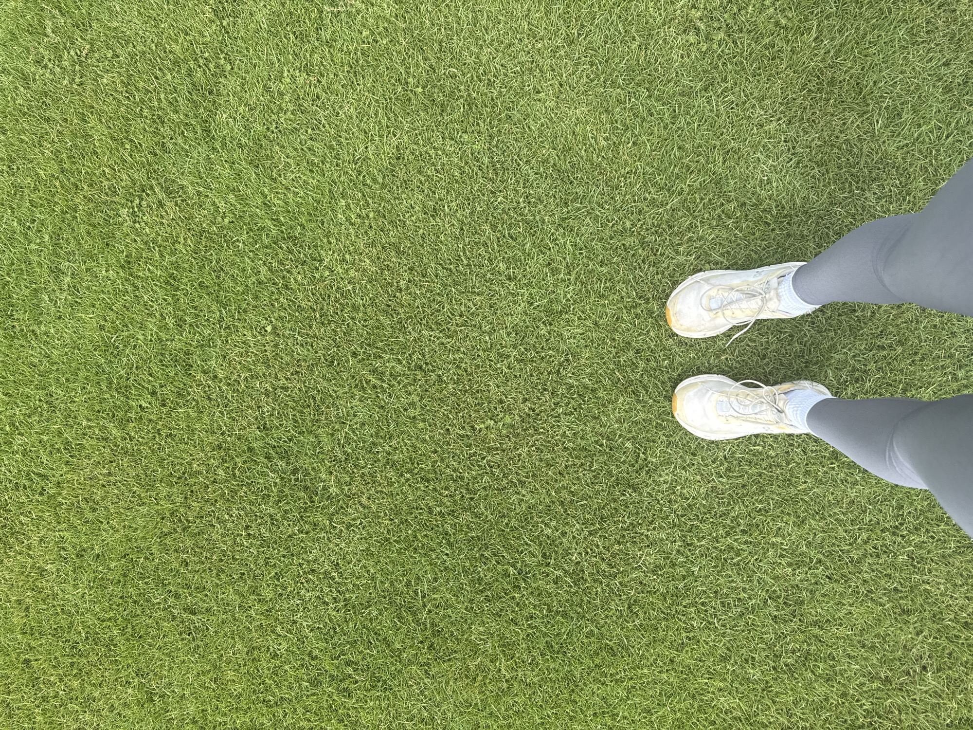 An aerial view captures a person's lower legs and feet dressed in athletic attire standing on a vibrant green lawn. The individual is wearing grey leggings and white athletic shoes with light blue socks. The lawn appears to be well-maintained and evenly cut, suggesting a park or a sports field. The scene is devoid of any urban elements, and there is no indication of the city of Miami, United States of America.