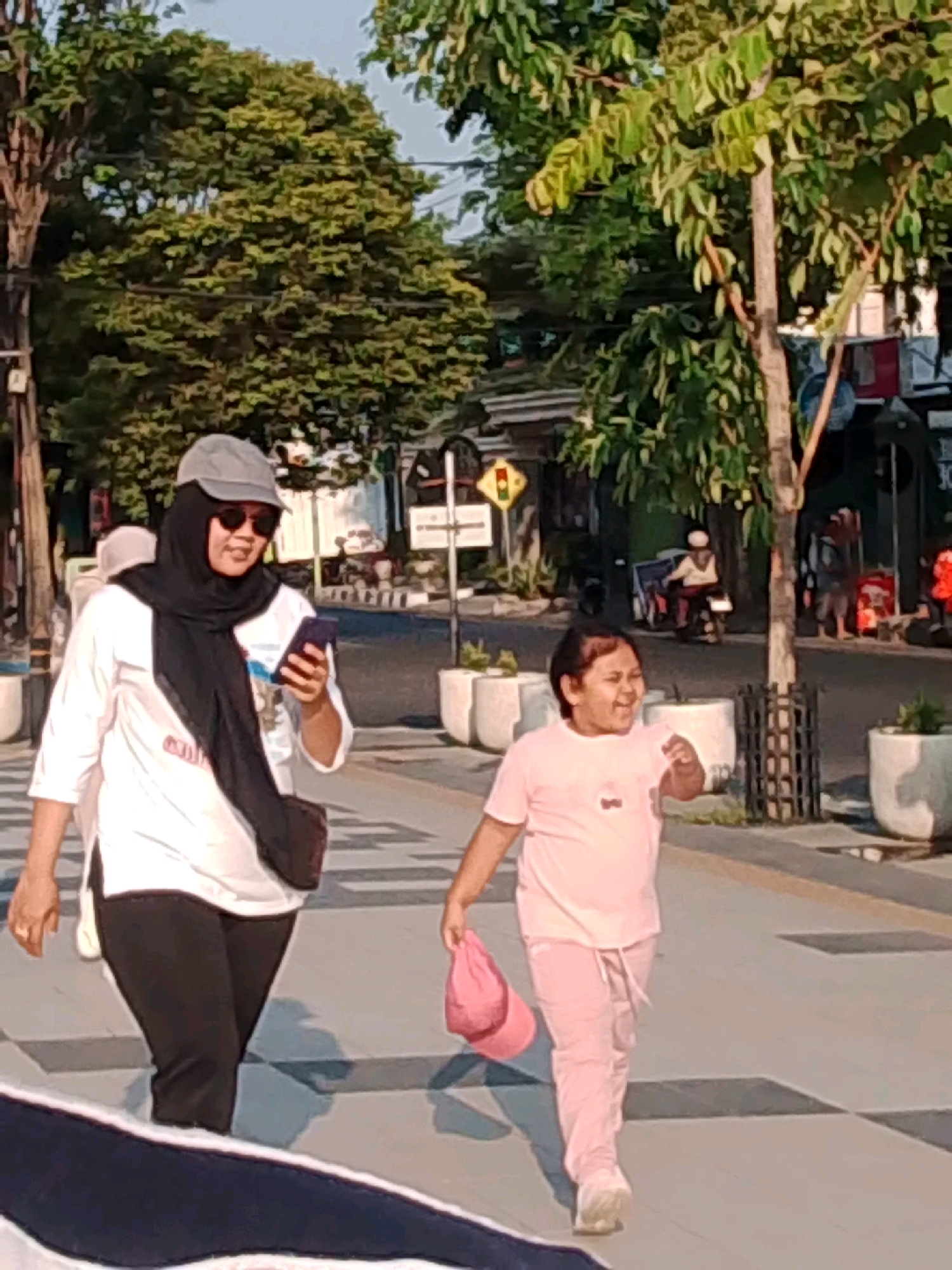 The image shows a street scene in Probolinggo, Indonesia. Two people are walking on a pedestrian walkway. On the left, a woman wearing a black hijab, sunglasses, and a white top walks while looking at her phone. She is also wearing black pants and a dark bag is slung across her body. On the right, a child in a pink t-shirt and pink pants runs with a pink bag in their hand. The walkway has a checkered pattern. In the background, there are trees, buildings, and people on motorcycles. The lighting suggests it is either early morning or late afternoon.