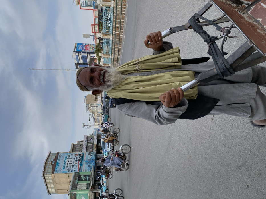 A male individual of advanced age, with a full white beard and a light brown skullcap, is depicted in the foreground, smiling and looking towards the camera. He wears a grey tunic (shalwar kameez), a dark vest, and a light olive green scarf draped over his shoulders. His hands grip the silver-colored metal handles of a two-wheeled, dark metal cart, with a reddish-brown wooden bed, which he appears to be pushing or pulling. The cart's frame is secured with black strapping material.
The scene unfolds on a paved street at what appears to be a busy urban intersection in Kandahar, Afghanistan. Numerous motorcycles are visible on the road, carrying one or more riders, some wearing traditional attire. Several cars are also present. To the left, a multi-story building with a light brown facade features large commercial signs with text in what appears to be Dari or Pashto, advertising electronics and other goods. Another blue-toned building with solar panels on its roof is visible further down the street to the right, also featuring commercial signage. A tall, slender antenna tower is discernible in the distance against a cloudy, overcast sky. The road surface is grey asphalt, and a light blue metal railing borders a median or raised pavement area to the right. Traffic signals and streetlights are also visible in the background.