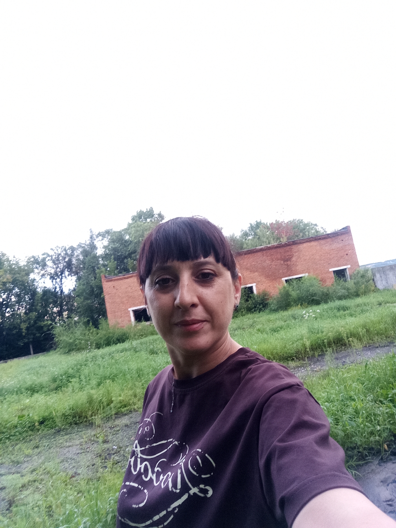 A woman with dark hair and bangs stands in front of an abandoned brick building. She is wearing a dark purple t-shirt with a white design. The building has broken windows and is surrounded by overgrown grass and trees. The sky is overcast. The location is the city of Belogorsk, Russia.