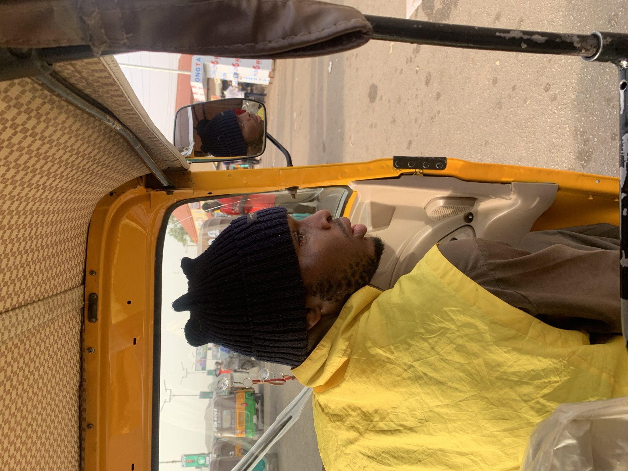 The image shows a man as a passenger inside a yellow motorized tricycle, commonly known as a keke napep, in Jos, Nigeria. The image is captured from an interior perspective, looking out and slightly to the right, with the man's profile prominent in the foreground.
The main subject is a man wearing a dark blue knitted beanie and a bright yellow long-sleeved garment, possibly a jacket or hooded top, over a darker, greyish-brown shirt. He has a neatly trimmed beard and is looking towards the right side of the frame, slightly upwards. A pair of glasses with red frames hangs around his neck or from his yellow garment, with the lenses visible near his chest. His reflection is also visible in the vehicle's side mirror, showing him wearing the same beanie and looking in a similar direction.
The interior of the tricycle features a yellow metal frame and a patterned roof lining with a brown and beige checkered design. Various screws and a small latch mechanism are visible on the interior side panel. The black metal frame of the vehicle's roof structure is at the top of the image.
Outside the tricycle, the setting appears to be a street or a busy area, possibly a market. Other tricycles, some green and yellow, are visible in the background. One tricycle clearly displays the alphanumeric text "PTA-6066" on its license plate. The background suggests a hazy or dusty atmosphere under natural daylight. A large white sign with partial text, including "ONGTA" and "R", is visible in the reflection of the side mirror, indicating a commercial establishment. The time of day appears to be daytime, with soft, bright light illuminating the scene. The overall impression is one of everyday street life and transport in Nigeria.