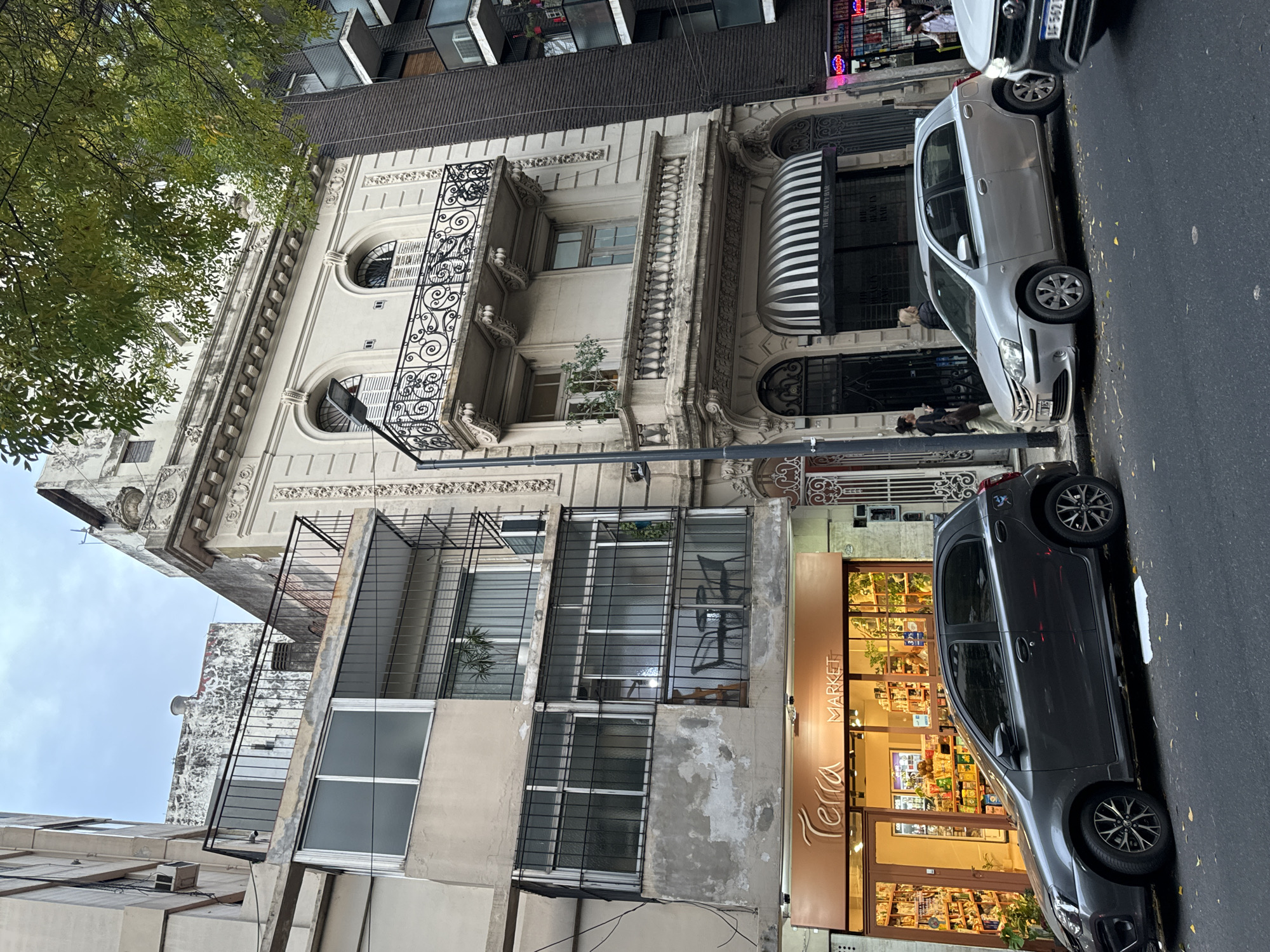 The image depicts a street scene in Buenos Aires, Argentina. On the right side of the frame, two parked cars, a silver hatchback and a dark grey compact, occupy the street lane. Adjacent to the grey car is a commercial establishment named "Terra Market," its storefront illuminated with warm light, displaying products on shelves inside.
The architecture of the buildings is varied. A multi-story structure features a classical facade with decorative mouldings, wrought iron balconies, and arched windows. Adjacent to this is a more modern building with a series of metal-framed windows.
Several individuals are present in the scene, standing and walking on the sidewalk in front of the buildings. The weather appears overcast, indicated by the soft, diffused light and the colour of the sky.