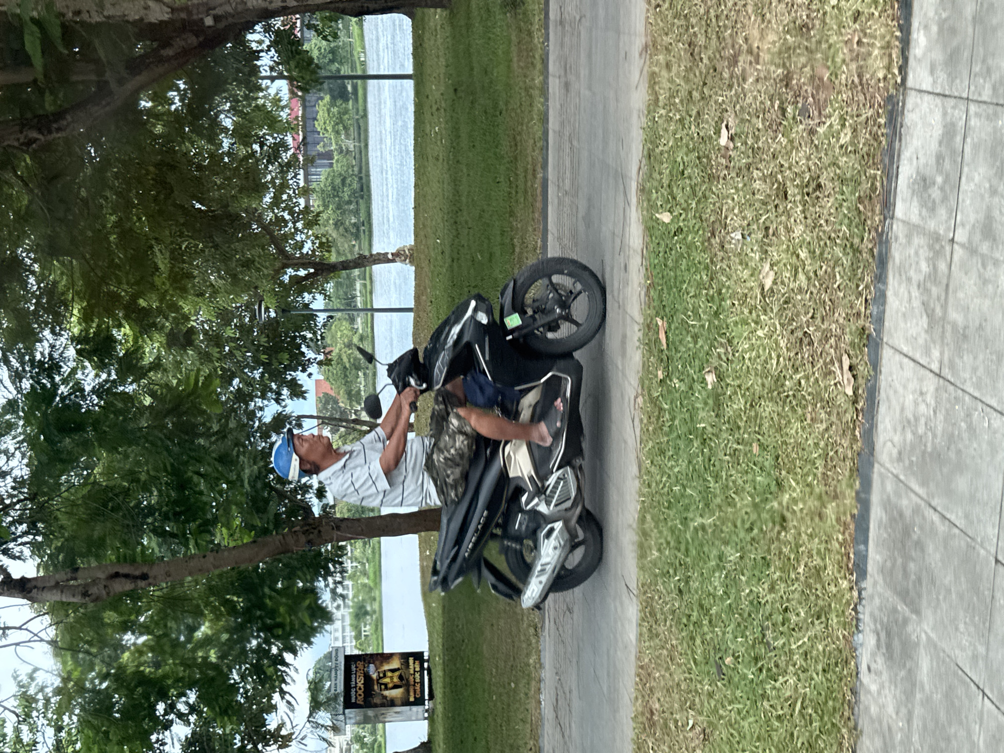 A middle-aged man wearing a blue helmet, a striped t-shirt, and camouflage shorts rides a black motorcycle on a paved path in a park. The man is facing right and appears to be looking ahead. The motorcycle has silver accents and a license plate. Behind the man, there is a body of water, trees, and a building with red roofs in the distance. A billboard for "Rockstar" is visible on the left side of the image. The ground is covered with grass and paved tiles. The scene suggests a casual outdoor setting in Phú Xuân Ward, Vietnam, likely a park or recreational area alongside a body of water.