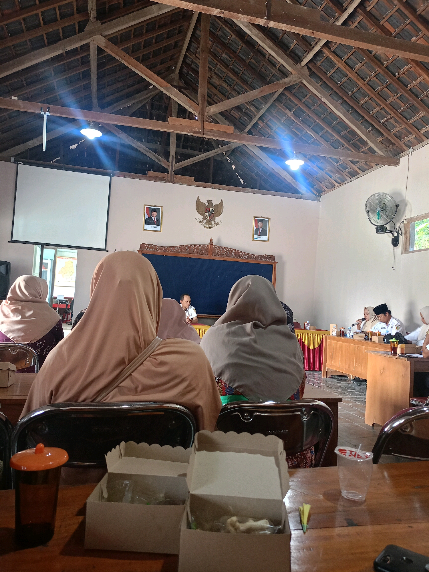 The image depicts an indoor meeting or presentation taking place in a room with a high, wooden-beamed ceiling. The room appears to be a community hall or a simple meeting space, possibly in the city of Kembang, Indonesia, given the cultural elements visible.
In the foreground, a wooden table is laden with several items. There are three open cardboard boxes, one containing some food items wrapped in plastic. To the right of these boxes, a partially filled clear plastic cup with a red-and-white striped straw is visible. A small orange-lidded dark amber glass is also present on the table, positioned to the left. Two chairs are visible in the lower half of the frame, with shiny chrome backs.
The middle ground of the image shows several people seated, facing a long wooden table at the front of the room. The majority of these individuals are women wearing headscarves, with their backs to the camera, suggesting they are attending the event. Some are seated in chairs, while others are behind the table.
At the front of the room, a long table is set up with various items, including bottles of water and a microphone. Several people are seated at this table, appearing to be speakers or organizers. One individual, a man wearing a white shirt and a black peci (a brimless cap common in Indonesia), is speaking into the microphone. Another person, also wearing a headscarf, is seated next to him, seemingly participating in the discussion. A man in a white shirt and dark pants is seated at the far end of the table, looking towards the front.
The walls of the room are a light color. On the back wall, behind the speakers, there is a large dark blue board or screen. To the left of the board, a framed portrait of a man in a suit is hung, and to the right, a coat of arms is displayed. A large projection screen is visible on the far left wall, suggesting a presentation is either ongoing or about to begin. A modern electric fan is mounted on the right wall, near a window that offers a glimpse of greenery outside. The overall atmosphere suggests a formal or semi-formal gathering.