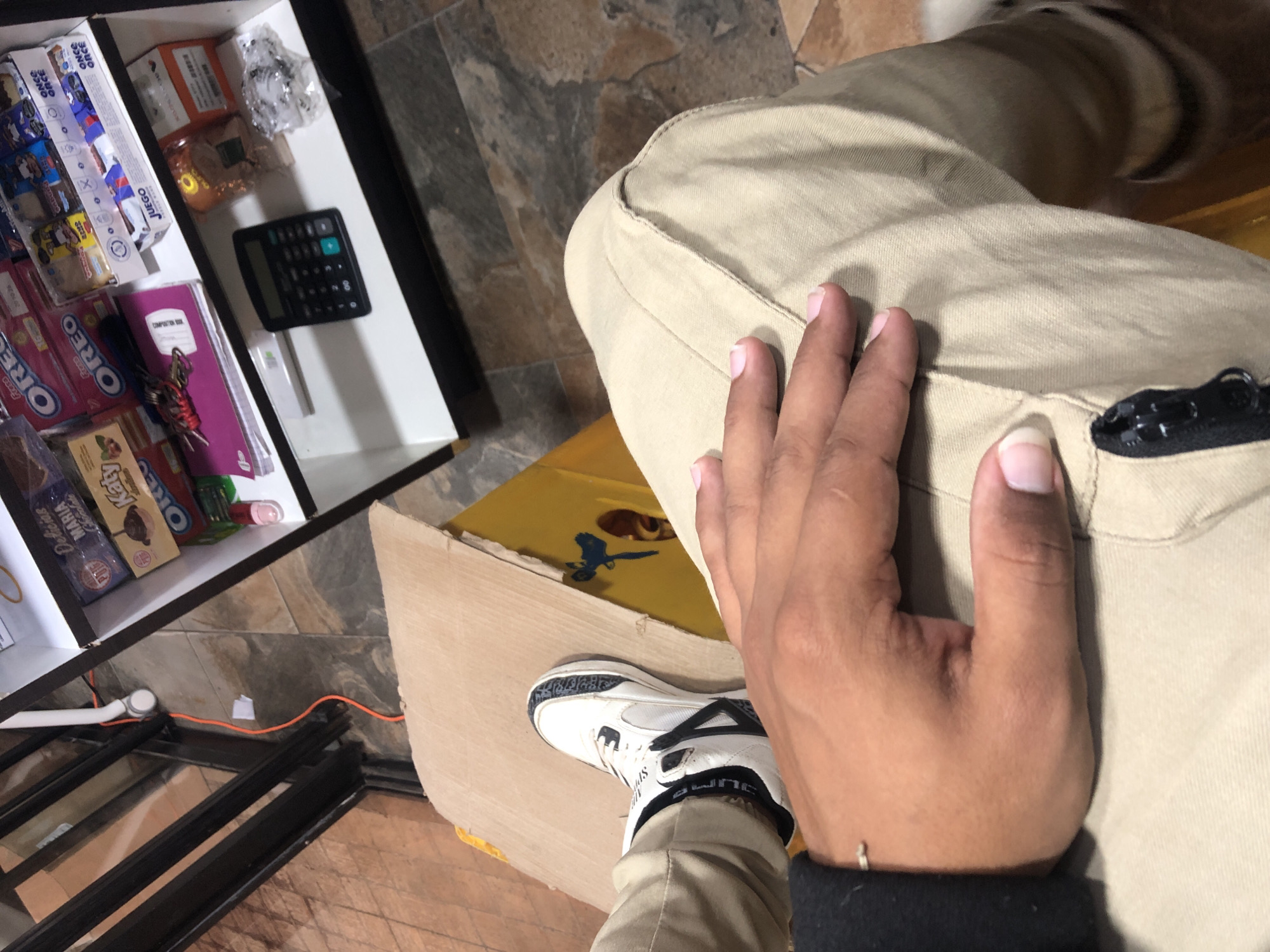 This eye-level shot captures a partial view of a person seated indoors in Juan Griego, Venezuela, with a focus on their right hand, leg, and foot, and an open drawer or shelf unit to their left. The person has a medium-brown skin tone, with their right hand resting casually on their thigh, which is clad in light khaki or tan pants. They are wearing a white and black sneaker with the brand "SPORTSLINE" visible on the side and a dark cuff of a long-sleeved shirt or jacket around their wrist.
To the left, an open, multi-compartment drawer or shelf is filled with various items. Prominently featured are numerous snack packages, including "OREO Freso" and regular "OREO" cookies, "Delicias MARIA" cookies, "Katy" brand items, and several "ONCE" brand packages, some labeled "JUEGO." Other packaged goods are also visible. A black calculator rests in one compartment, alongside a purple "COMPOSITION" notebook and a set of keys tied with a red string.
The setting features a tiled floor with irregular gray and brown patterns. Below the drawer unit, an orange electrical cord runs along the floor next to a white conduit and a black metal railing or structure. Part of a yellow surface, possibly a chair or table, is seen beneath the person's leg, next to a light-colored cardboard box. The overall impression is a casual, possibly domestic or small retail environment, with the various snack items suggesting a place where goods are sold or stored for consumption. The lighting suggests it is daytime.