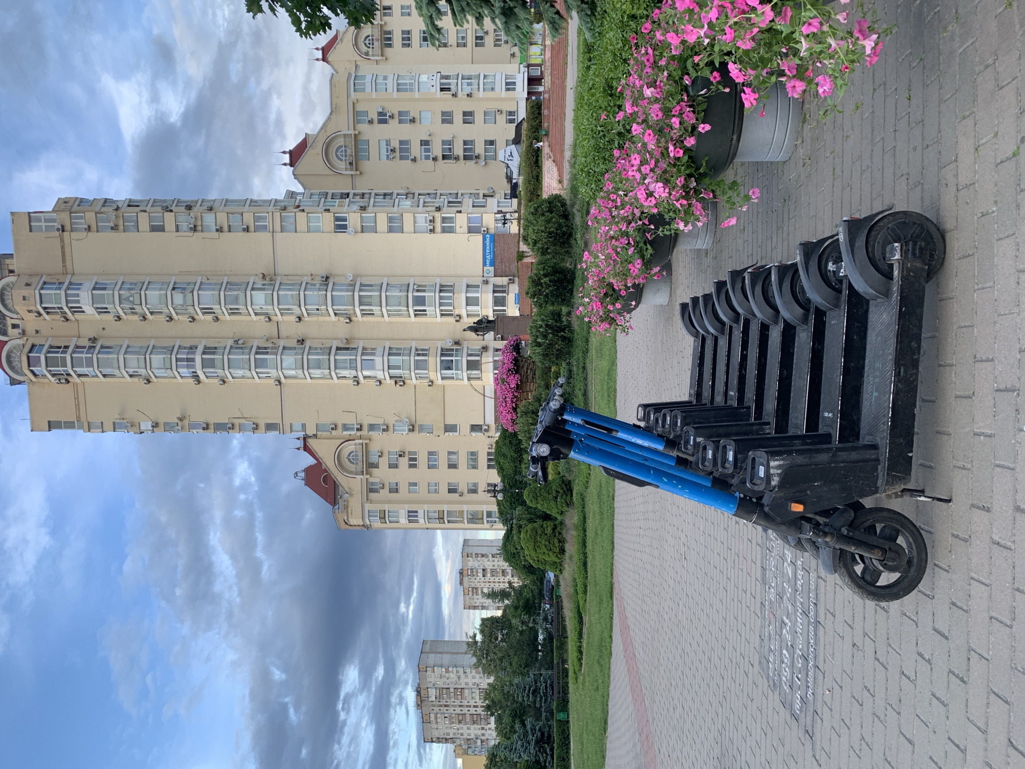 The image depicts a row of parked electric scooters on a brick sidewalk in Kyiv, Ukraine. The scooters are mostly black, with blue handlebars on the visible ones. To the right, flower pots filled with pink petunias line the sidewalk. Behind the scooters, there's a green lawn area with hedges and trees, separating the sidewalk from buildings in the background. The main building in view is a multi-story apartment complex with light-colored walls and numerous windows. A smaller, older building can be seen further back. The sky is overcast with varying shades of blue and gray clouds. A sign is visible on one of the buildings in Ukrainian. The text is “450 24 Сталинграда, пр. Г. Сталинграда, 14”.
