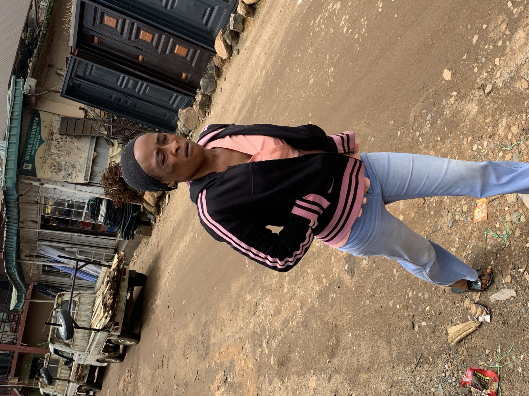 A woman wearing a black and pink tracksuit jacket, a pink shirt, and light blue jeans walks on a dirt road in Gura Top, Nigeria. She is also wearing black sandals. In the background, there are two white pickup trucks loaded with lumber and a spare tire. Adjacent to the trucks is a building with various items displayed outside, including what appear to be metal doors and coiled wires. The ground is covered with dirt and scattered debris.