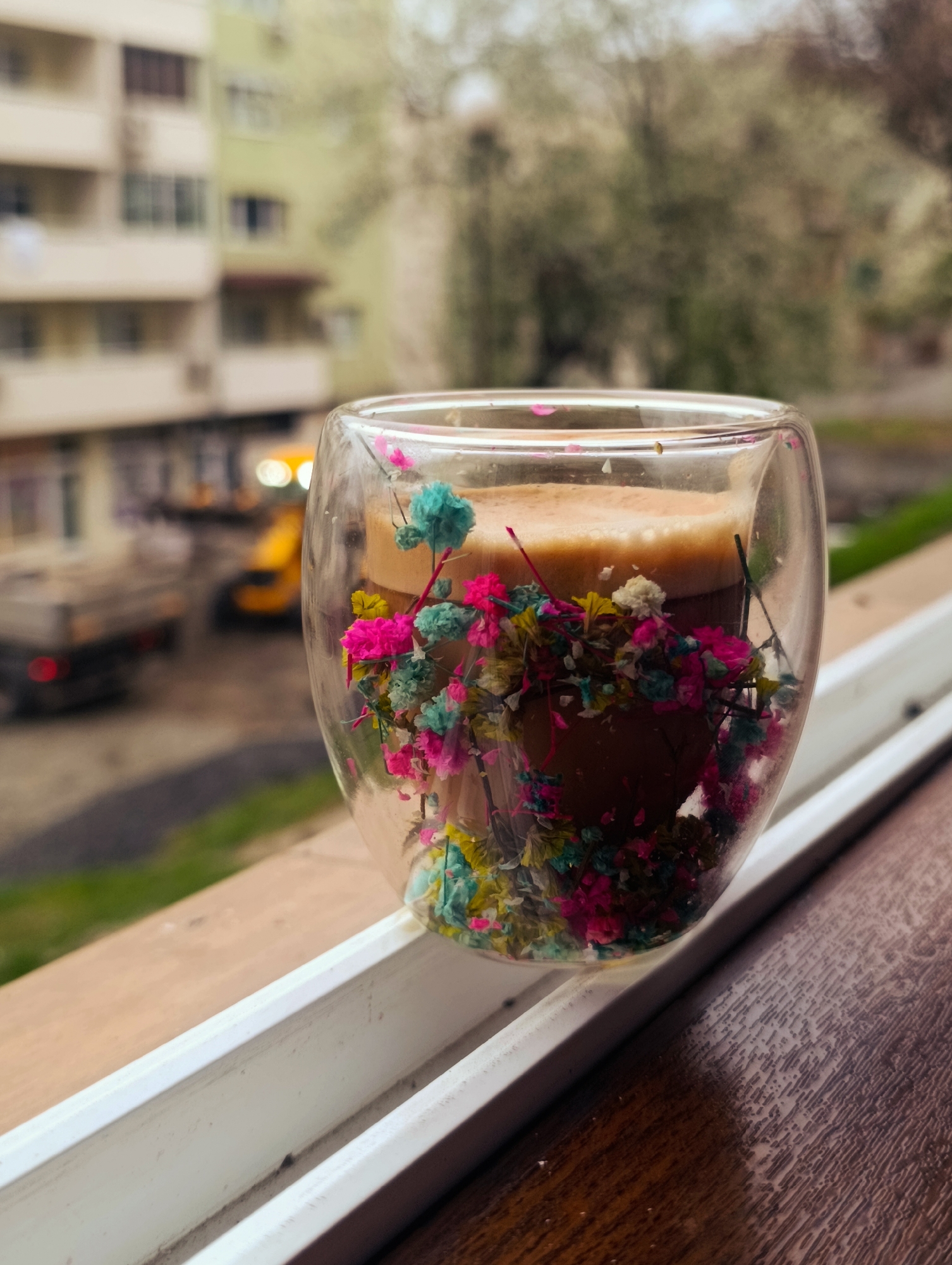 A glass cup is the primary subject, filled with coffee and decorated with vibrant, small, dried flowers in shades of pink, blue, and yellow. The cup is placed on a white, wooden window ledge that transitions into a brown, wooden surface.
Through the transparent glass, the background reveals an urban scene. Several buildings, painted in light shades, are visible across the street. A yellow construction vehicle and a truck can be seen on the road, suggesting possible construction or road work activity. The location is likely in Kardzhali, Bulgaria. The image evokes a serene and casual atmosphere.