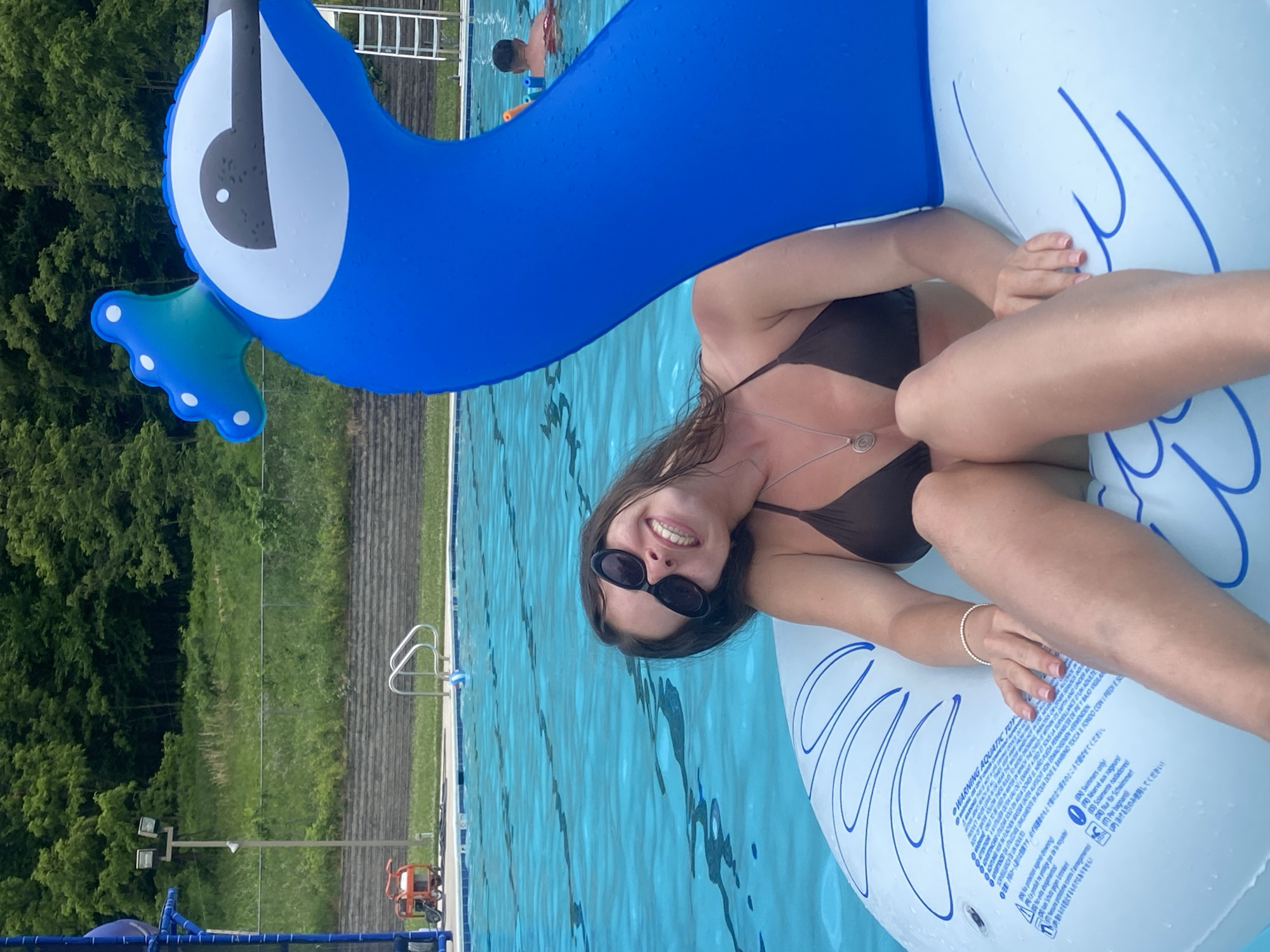 A woman in a brown bikini is floating on a large inflatable pool toy that resembles a blue bird's head and neck. She is wearing dark sunglasses and has a necklace with a spiral pendant. The inflatable has a large white and blue section with text warning about its use. The woman is smiling and looking at the camera. She is in a swimming pool with clear blue water. In the background, there are trees and a pool ladder. Another person, a child, is visible in the pool in the distance. The overall scene suggests a sunny day at an outdoor swimming pool. The text on the inflatable toy provides safety warnings in multiple languages, including English, French, Italian, Spanish, German, and Japanese. The location is identified as Washington Township, United States of America.