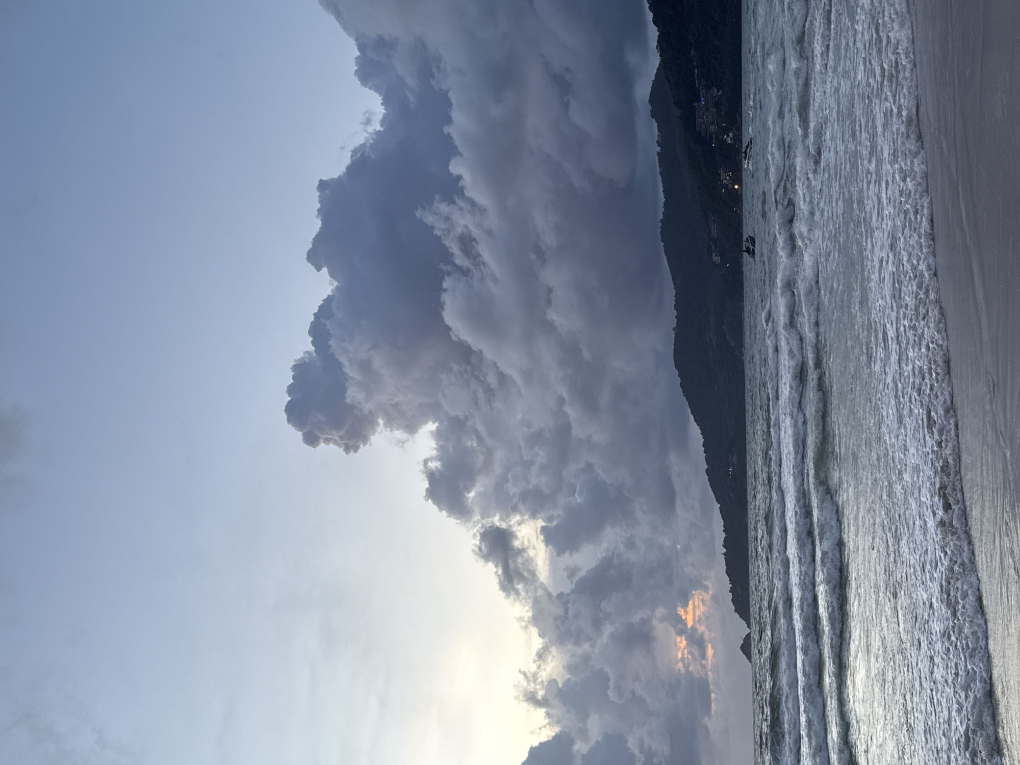 This is a medium shot of a beach in Patong, Thailand during a cloudy sunset. The sky is filled with dark, dramatic clouds, with a sliver of orange light peeking through on the left. The ocean has moderate waves crashing onto the shore, creating white foam. In the distance, a lush, green hill rises from the water, dotted with buildings and lights. Two small boats are visible on the water, one of which appears to be moving at speed. The sand in the foreground is wet and reflects the dim light.