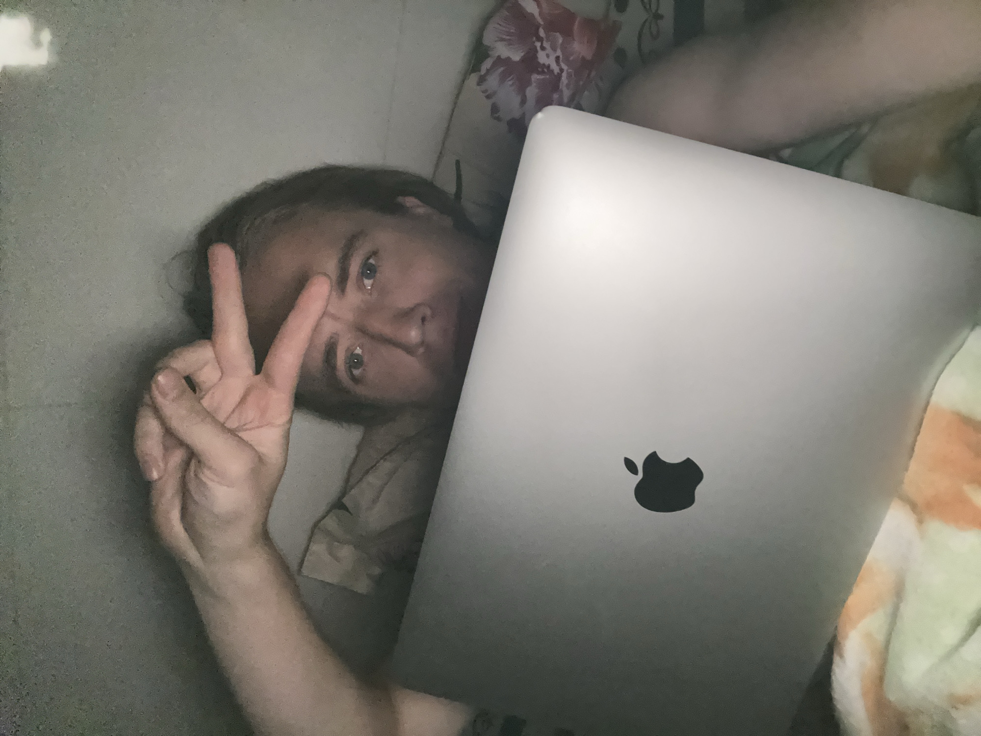 The image depicts a person lying down and holding a silver laptop with an Apple logo. The person is looking at the camera and has their right hand raised, with two fingers extended in a peace sign gesture. The person appears to be in a bedroom, with bedding visible in the background, including a pillow with a floral pattern. The lighting suggests it is nighttime or a dimly lit room. There is no information in the image that can provide the location in the city of An Hữu, Vietnam.