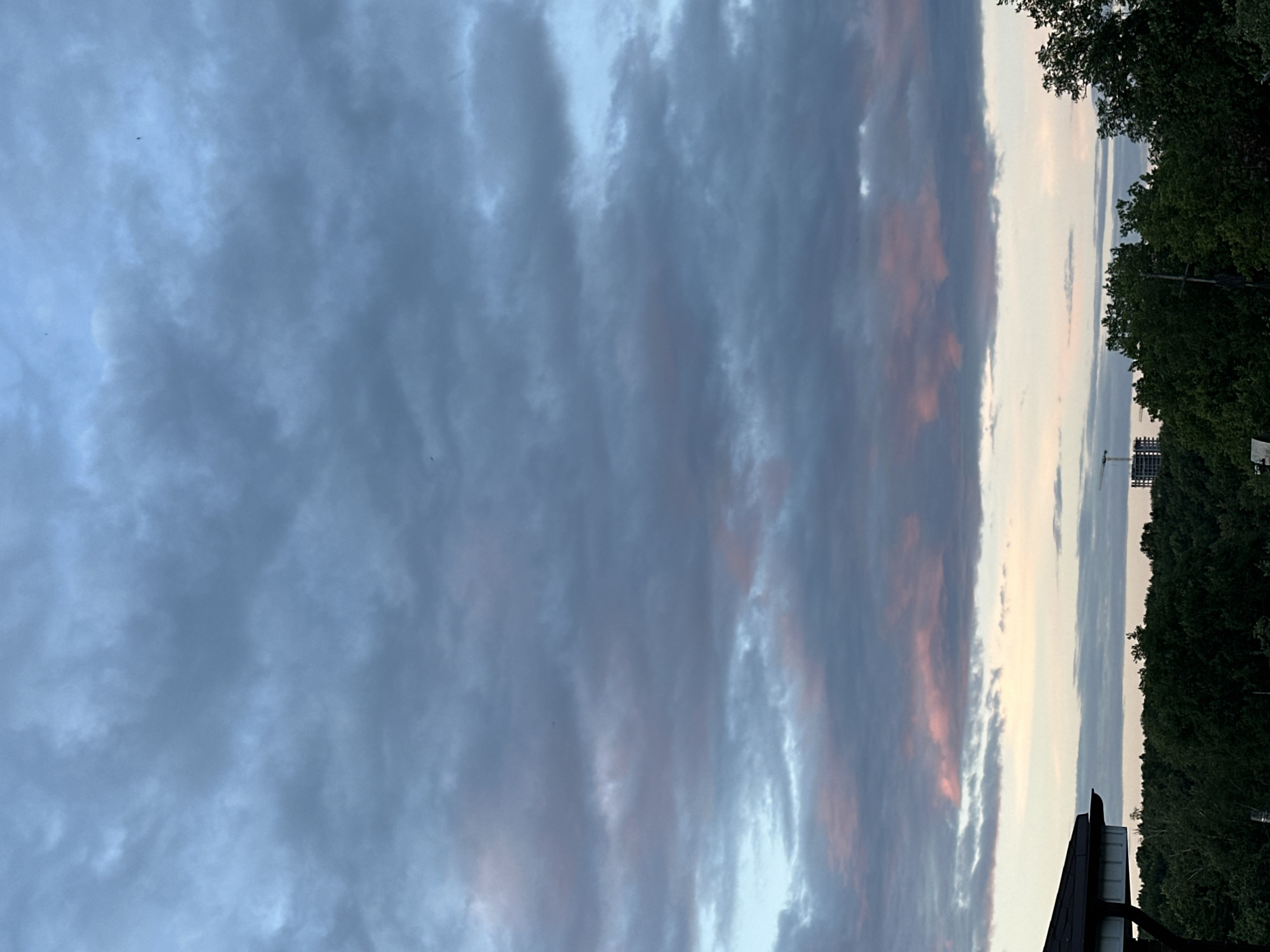 The image depicts a twilight scene over a densely wooded area, possibly a park or the outskirts of a city. The sky is filled with a dramatic display of clouds, predominantly dark grey with streaks of soft pink and pale blue, suggesting the transition from day to night or an impending storm. In the distance, silhouetted against the horizon, are two construction sites, each featuring a tall, unfinished building and a prominent construction crane. A portion of a dark-colored roofline with white trim is visible in the lower left corner, indicating a structure close to the viewpoint. The overall impression is one of a natural landscape juxtaposed with urban development. The location is identified as Nizhny Novgorod, Russia. There are no people visible in the image.