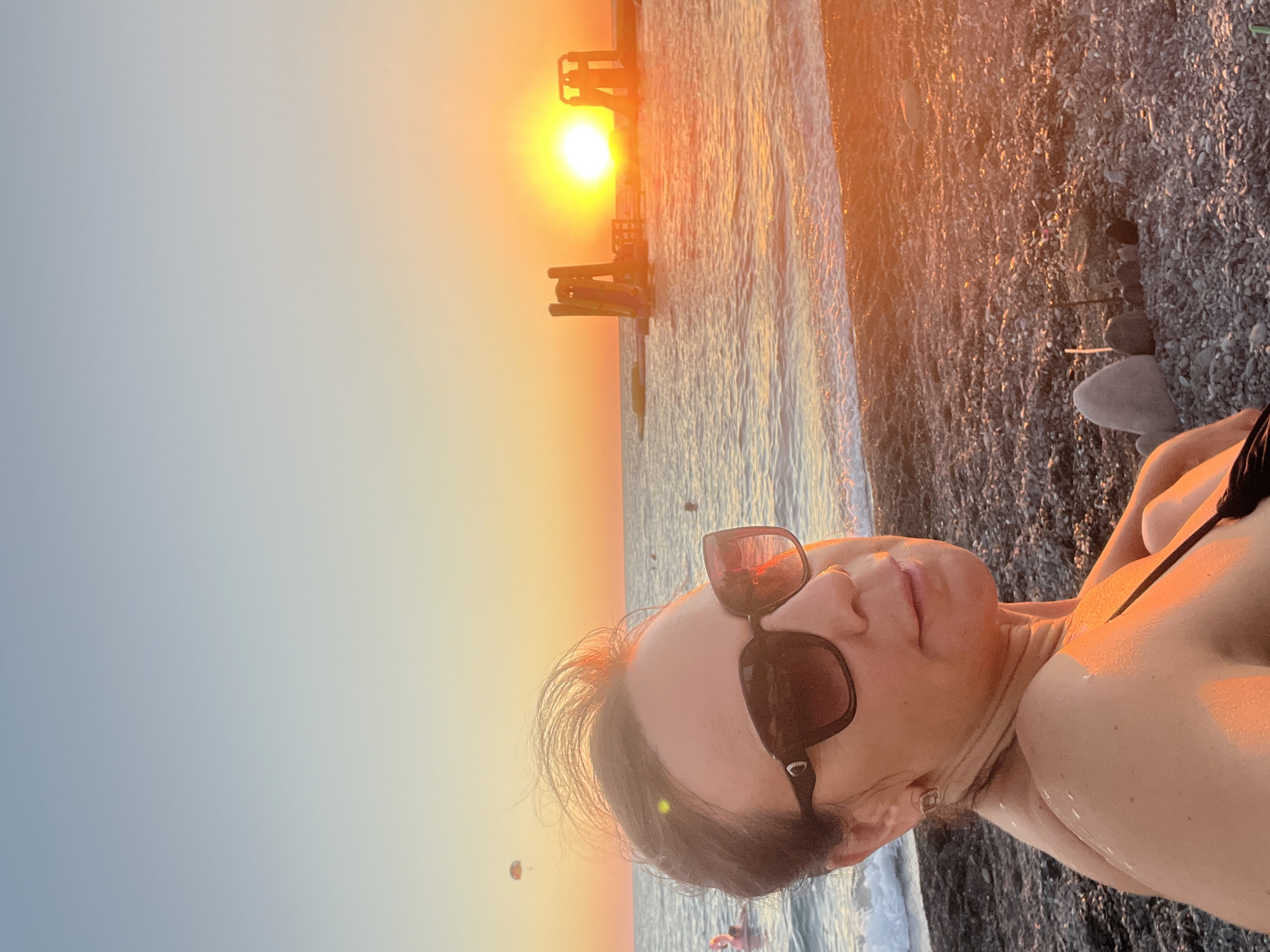 An adult female is shown in a close-up selfie on a pebbled beach in Sochi, Russia, at sunset. She is wearing large, dark sunglasses and a black bikini top. The sun is low on the horizon, casting a warm, orange glow across the sky and water. In the background, the Black Sea is visible, with gentle waves rolling towards the shore. A floating water park with slides is present in the water to the right, and a paraglider can be seen in the sky to the left. The beach is covered in small, smooth pebbles. The overall impression is one of a peaceful evening at a seaside resort.