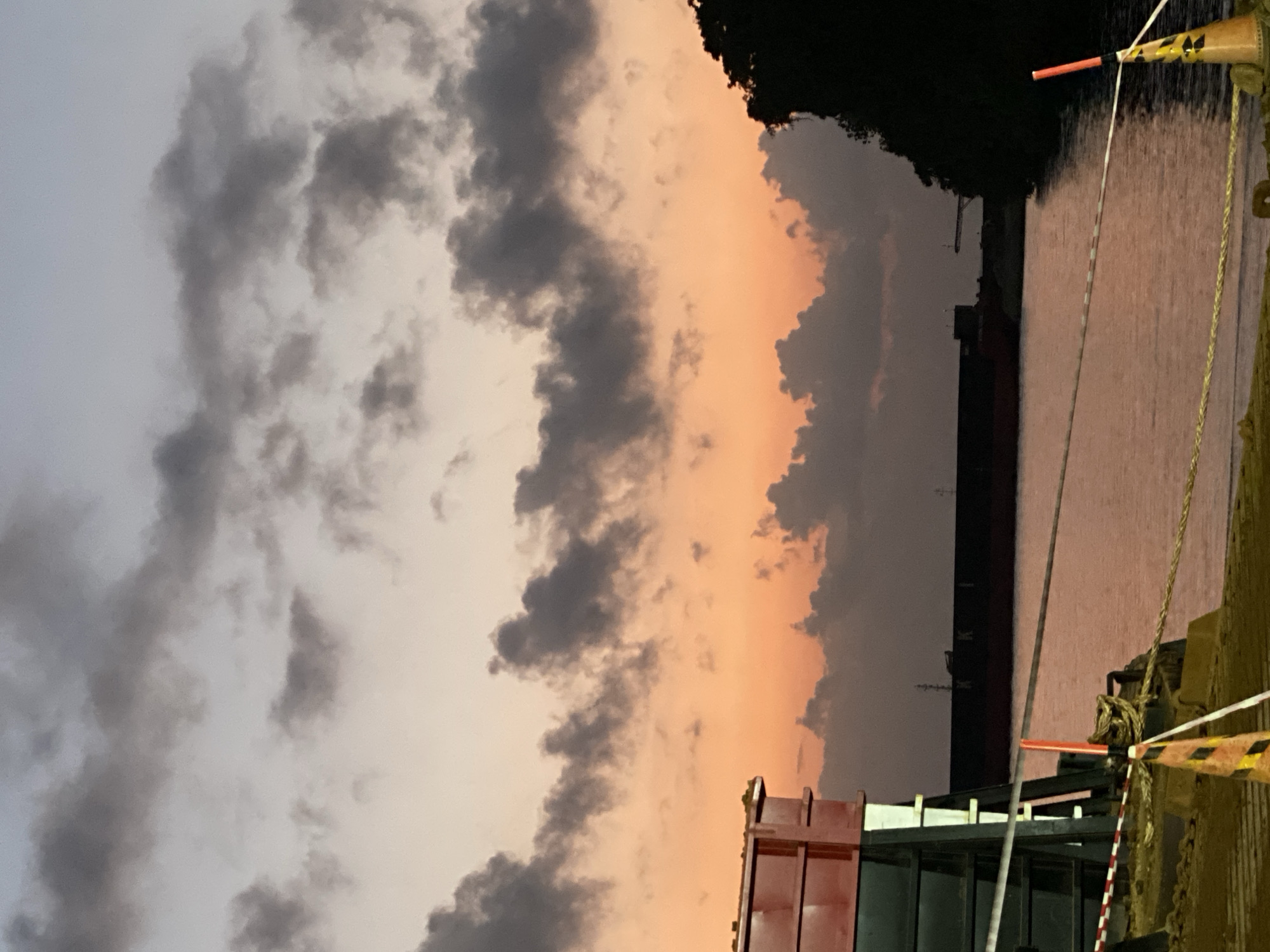 A twilight scene unfolds over a body of water, possibly a harbor, in Sambalagi, Indonesia. The sky is a gradient of soft blues and oranges, streaked with dark, textured clouds. In the foreground, a red and metallic structure, possibly a cargo container or part of a dock, occupies the left side of the frame. Safety cones and ropes are visible, suggesting a designated area or construction zone. Across the water, a large, dark ship is silhouetted against the fading light, with some indistinct markings visible on its hull. The water reflects the muted colors of the sky, creating a calm and serene atmosphere. The right edge of the image is dominated by the dark silhouette of dense foliage, indicating a natural landscape bordering the urban or industrial setting.