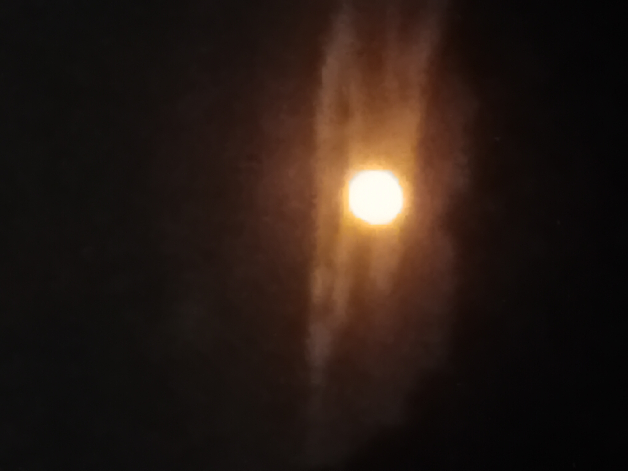 A full moon shines through patchy clouds, casting a warm glow on the scene. The moon is centered in the frame, appearing as a bright, luminous orb. The surrounding clouds are wispy and semi-transparent, allowing the moonlight to filter through. The overall atmosphere is dark and slightly obscured, typical of a nighttime sky. No specific details about Verkhniachka, Ukraine, are discernible from the image itself. There are no people, objects, or actions depicted.