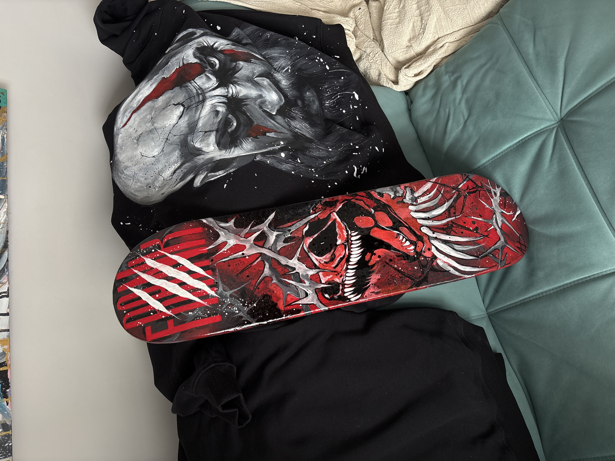 A black hoodie and a custom-painted skateboard, both featuring dark and aggressive artistic designs, are displayed on a bed or sofa. The arrangement highlights two distinct art pieces in an indoor setting.
The black hoodie, serving as the base, showcases a large, detailed painting of a monstrous or demonic head on its front. The head is rendered in shades of white, grey, and black, with striking red accents resembling deep scars or gashes across its face and around the eyes. The style incorporates splatters and sharp lines, giving it a gritty, distressed look.
Resting diagonally on top of the hoodie is a skateboard, painted predominantly in red and black with white details. Its design features a skeletal or monstrous mouth with sharp white teeth, surrounded by ribcage-like structures and abstract, thorny patterns. The left end of the skateboard displays three prominent white claw marks over a background of red stripes, below which is some stylized, partially obscured red text. White paint splatters are scattered across the skateboard's surface, consistent with the hoodie's aesthetic.
The items are laid over a textured, light beige blanket or throw on the upper right, and a quilted turquoise-blue surface, likely part of a sofa or bedding, on the right and bottom. A plain white wall is visible on the left side of the frame, with a sliver of another object, possibly a canvas with colorful paint splatters, visible at the far left edge. The scene suggests a presentation of custom-made, dark-themed artwork.