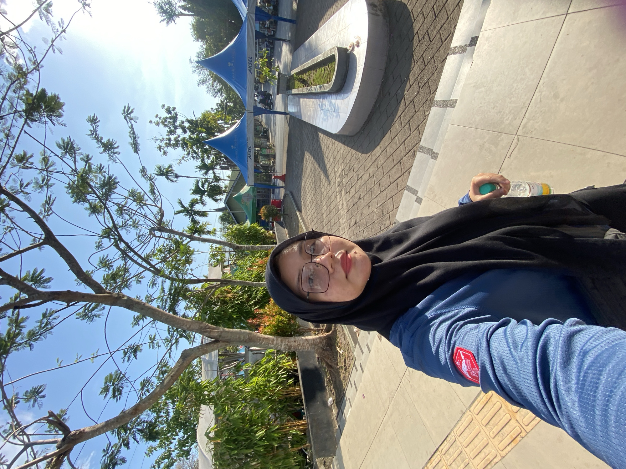 A young woman wearing a black hijab and glasses takes a selfie outdoors in Banjarmasin, Indonesia. She is wearing a blue long-sleeved shirt with a red and white emblem on the left sleeve. In her right hand, she holds a clear plastic bottle of water with a green cap. The background shows a paved area with trees, some green tents, and other structures. The sun is shining brightly, casting shadows. The environment appears to be an open public space within the city, possibly a park or a marketplace, with a clear blue sky overhead.