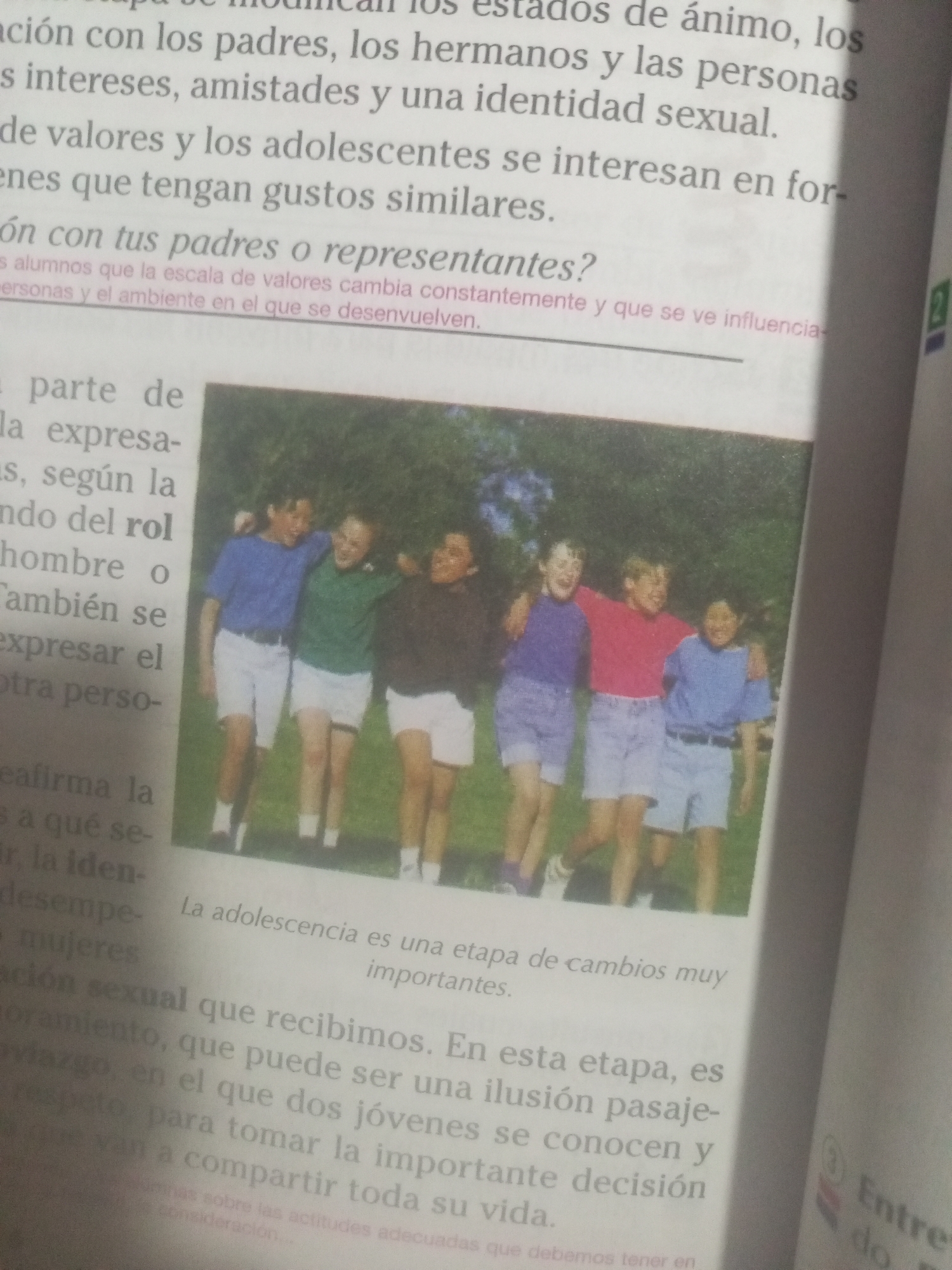 The image shows an open book. The left page displays text with a heading that reads "La adolescencia es una etapa de cambios muy importantes." The text discusses how adolescence is a stage of important changes, including relationships with parents, siblings, and other people, as well as the development of interests, friendships, and sexual identity. It also mentions that values and adolescents' interests change and that they look for similar tastes. The right page has text and a section that appears to be a continuation of the topic.
Below the text on the left page, there is a photograph featuring four young people walking with their arms around each other's shoulders, appearing to be friends. They are all wearing casual clothing, including t-shirts and shorts. The background of the photograph appears to be an outdoor setting with greenery.
The image does not contain any information that can be used to locate it in La Tagua, Venezuela.
