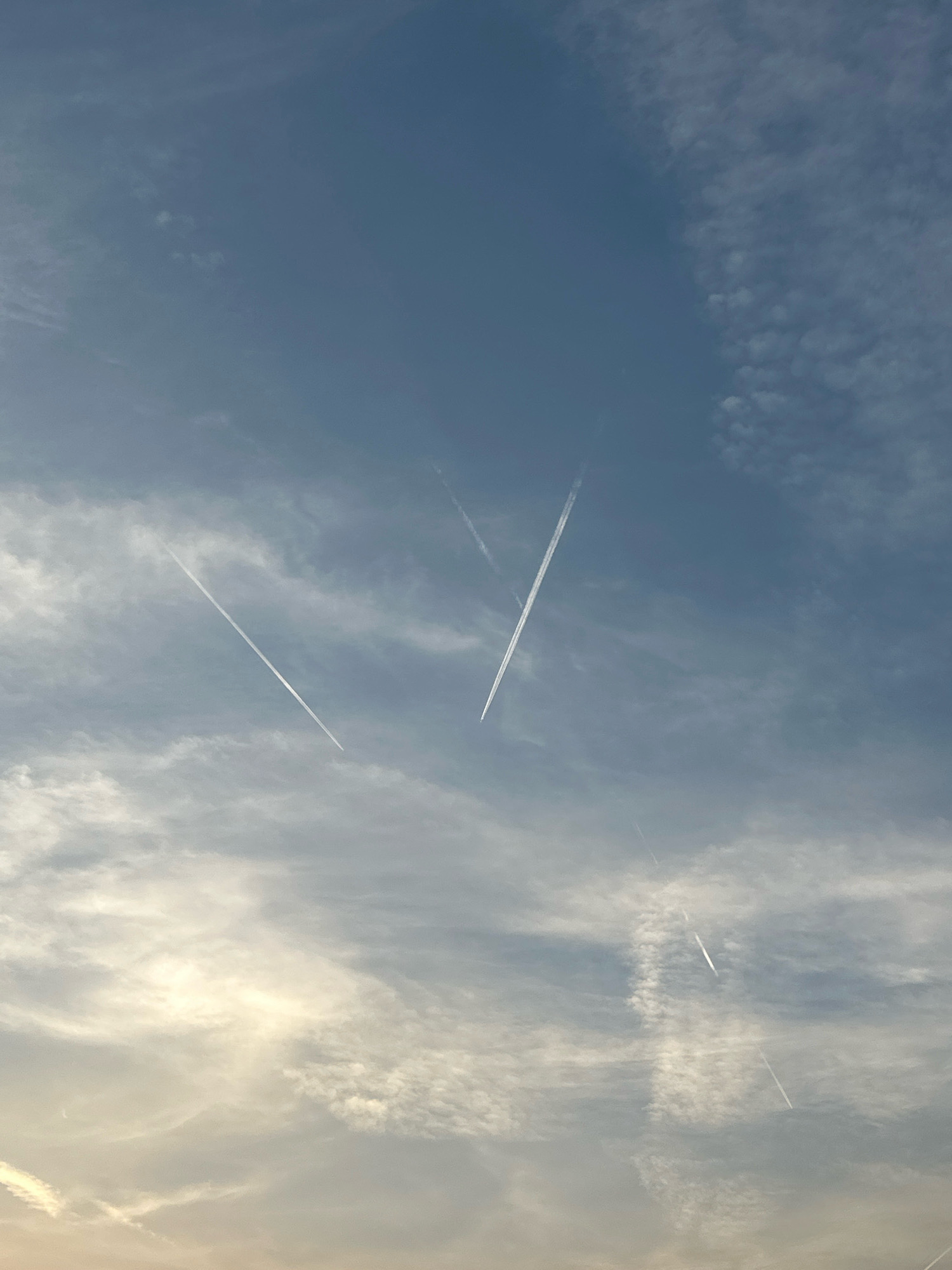 This is a vertical, medium-shot photograph of the sky above Buljarica, Montenegro, featuring contrails from airplanes and scattered clouds. The sky is a gradient of blue, lighter towards the bottom where it is illuminated by sunlight, and darker towards the top. The clouds are wispy and appear in various densities, with some areas being more opaque and others thinly spread. Multiple white contrails are visible, streaking across the sky at different angles. One prominent contrail forms a "V" shape in the upper middle section of the image. Another long, straight contrail runs diagonally from the middle left to the bottom right. Several other fainter contrails are also present, some appearing to intersect. The overall impression is one of an active sky with aircraft traffic.