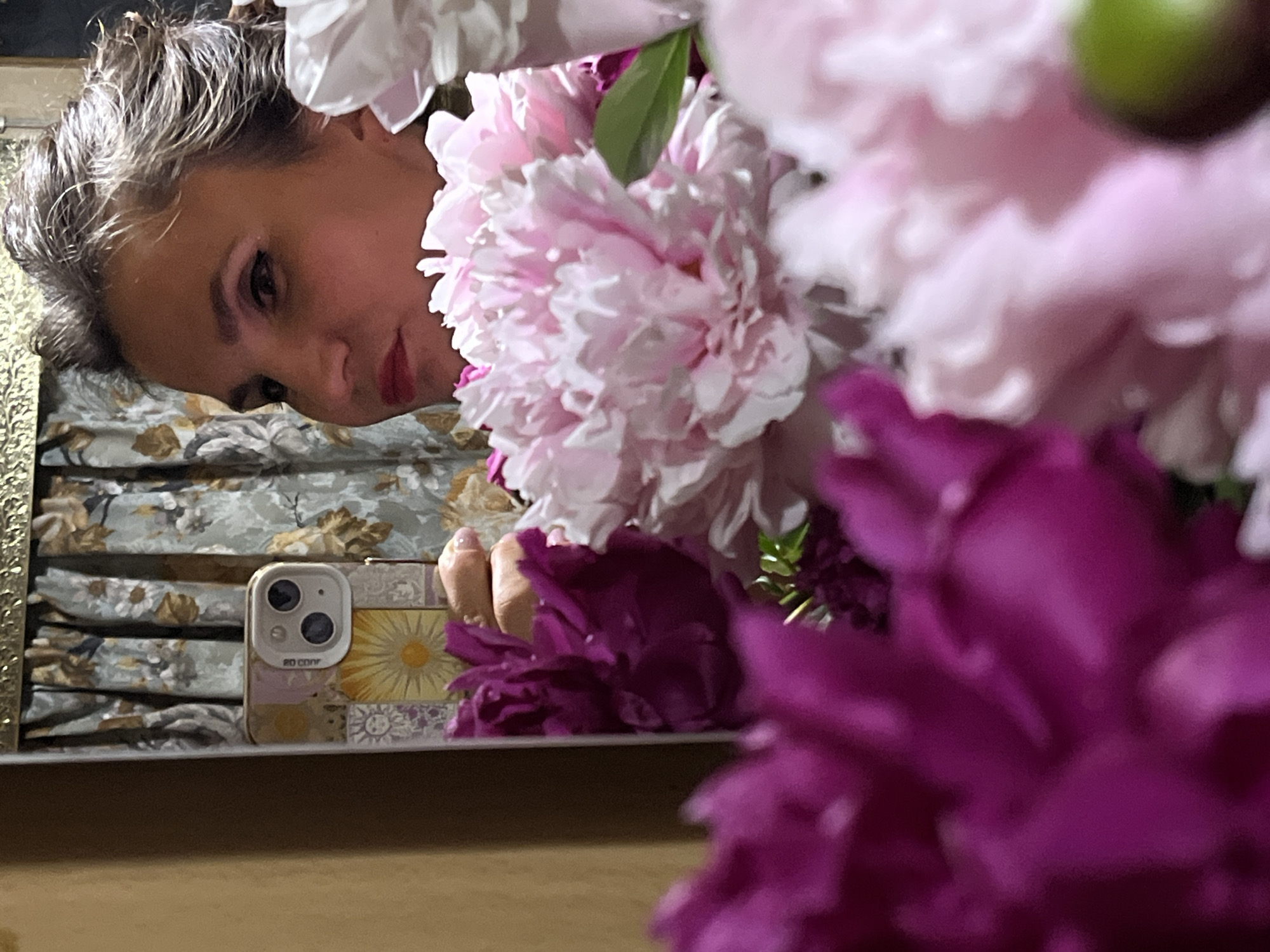 The image depicts a close-up reflection of a woman in a mirror. The woman is facing to the left, with her head slightly tilted. She has her hair pulled back and is wearing makeup, including lipstick and eyeliner. In the foreground, a bouquet of peonies is visible, with some flowers in shades of pink and deep purple. The background includes a patterned curtain and a part of the mirror's frame, which has a decorative, golden edge. A smartphone with a decorative case is held by a hand, partially obscuring the bouquet and the woman's reflection. The scene appears to be indoors. This image was taken in Petropavlovsk-Kamchatsky, Russia.