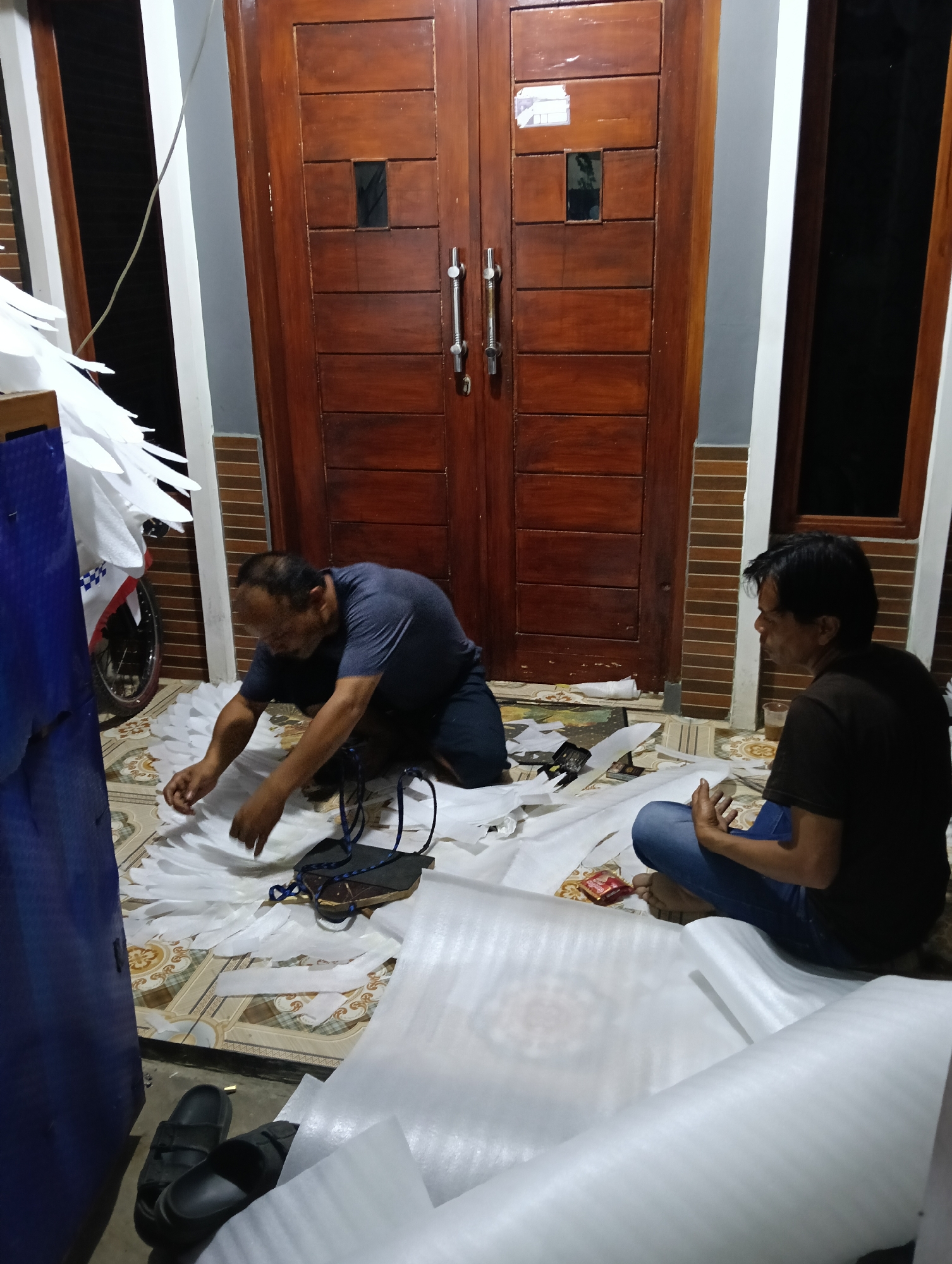 Two adult males are on the floor in front of a wooden double door. The male on the left is kneeling and appears to be working with white material that is spread across the floor. He is wearing a dark blue t-shirt and dark shorts. The male on the right is sitting cross-legged and looking towards the left. He is wearing a dark t-shirt and blue jeans. The floor is tiled with a patterned design. There are rolls of white material and scattered white pieces of material on the floor. To the left of the frame, part of a motorcycle is visible, with white feathered-like decorations attached to it. The environment appears to be an outdoor covered area, possibly a porch or overhang, with a house facade in the background. The lighting suggests it is evening or nighttime.