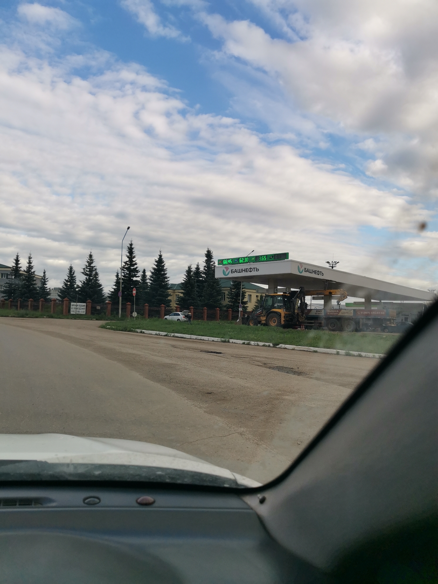 This image shows a gas station in Oktyabrsky, Russia, on an overcast day. A white car is parked on the side of the road near a row of evergreen trees and a fence. In the background, a yellow excavator and a truck are parked under the canopy of a Bashneft gas station. The gas price display shows "66.45 62.30 33.65". A sign in Russian, which can be partially translated as "RENT OF BUILDINGS AND PREMISES", is visible near the trees. The image is taken from inside a vehicle, with the dashboard visible in the foreground, suggesting the photographer is a driver. The sky is filled with clouds, indicating it's likely daytime.