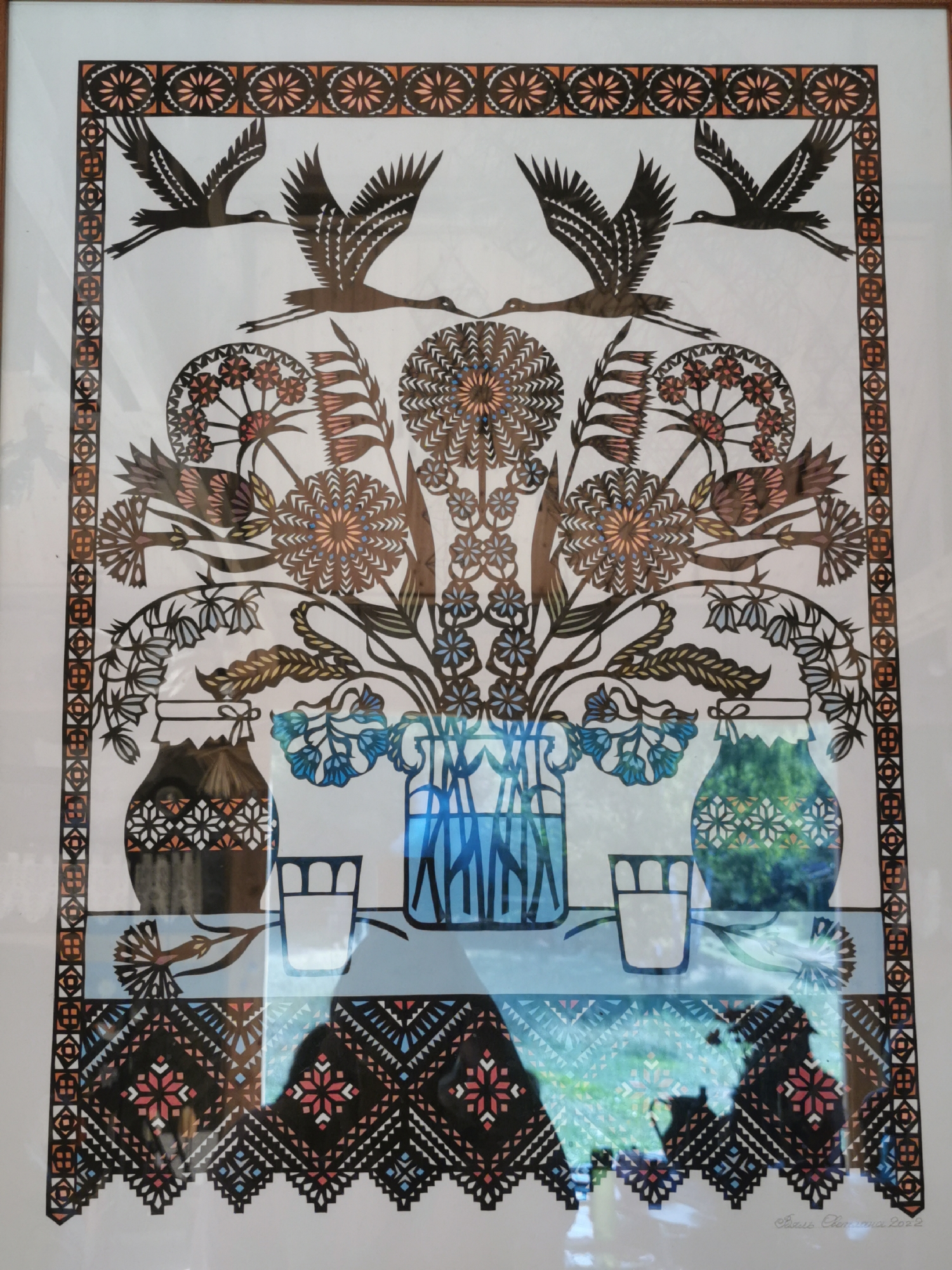The image displays a framed piece of folk art, likely a paper cut-out, with a symmetrical design. The artwork features several stylized elements, including four birds in flight at the top, two on each side, resembling cranes. Below the birds is an elaborate floral arrangement emanating from two vases and a pitcher. The floral motif is intricate, with circular, star-like flowers and delicate branching stems. Two glasses are placed on a table-like surface, situated below the vases. The entire composition is framed by decorative borders along the top, sides, and bottom, featuring geometric patterns and floral elements in black, orange, and white. The bottom border transitions into a more complex, zig-zagging pattern with diamond shapes in red, blue, and black. The artwork is signed in the lower right corner with Cyrillic script and the year "2022." The lighting suggests the artwork is indoors, possibly a home or gallery, with reflections on the glass of the frame indicating the presence of ambient light and possibly a window or other light source. The specific location within Шчарчоўскі сельскі Савет, Belarus is not discernible from the image itself.