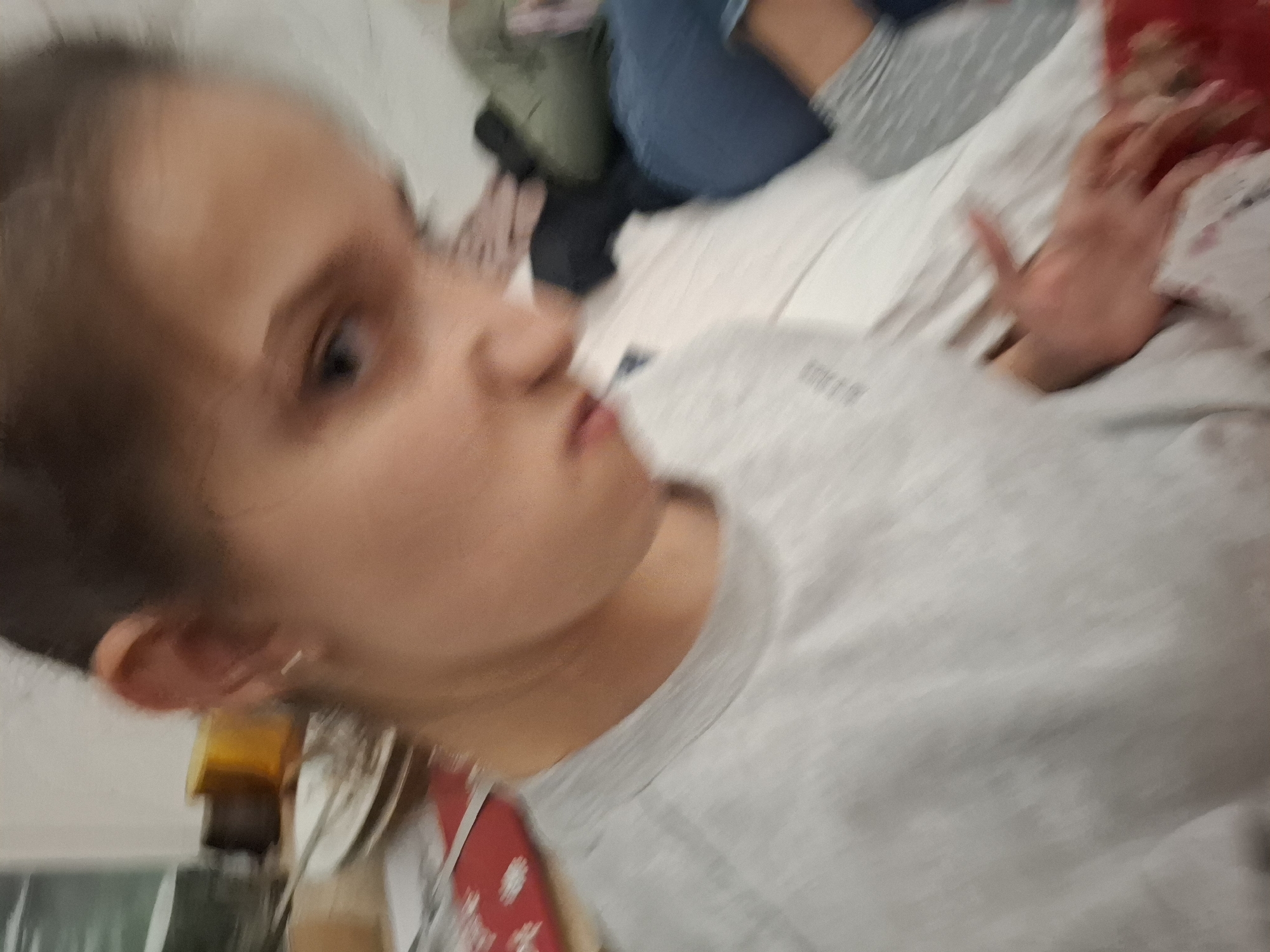 A young person with fair skin, wearing a gray long-sleeved shirt, is lying on a bed covered with white sheets. Part of another person's legs, wearing green pants and blue jeans, can be seen in the upper center of the frame. A hand, presumably belonging to the young person, is extended towards the upper right corner of the image. Some blurred objects are visible in the lower left corner. The photograph appears to be taken from a low angle, possibly with the camera close to the subject's face. The setting is unclear but presumed to be indoors based on the bed and clothing. The location in Panagyurishte, Bulgaria is not visibly confirmed.