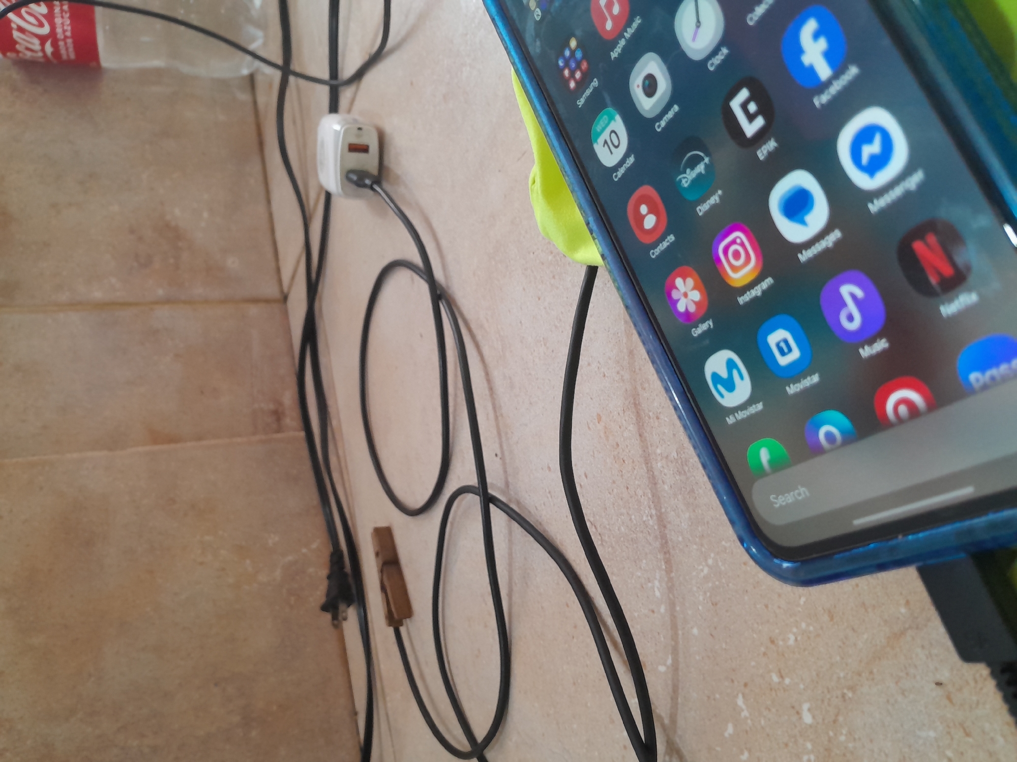 The image shows a close-up of a smartphone with a blue case, lying on a beige tiled surface. The phone screen is on, displaying a grid of app icons, including Samsung, Calendar, Contacts, Gallery, Instagram, Messages, Facebook, Music, and Netflix. A black charging cable is connected to the phone and extends out of frame. In the background, a white power adapter is plugged into an unseen outlet, with black wires running across the tiled surface. A wooden clothespin is visible on one of the black wires. Further in the background, a partially visible plastic bottle of Coca-Cola is present. The overall environment appears to be indoors, possibly a kitchen or countertop area. There is no identifiable presence of people or specific landmarks that would indicate a location in Santa Ana, Venezuela.