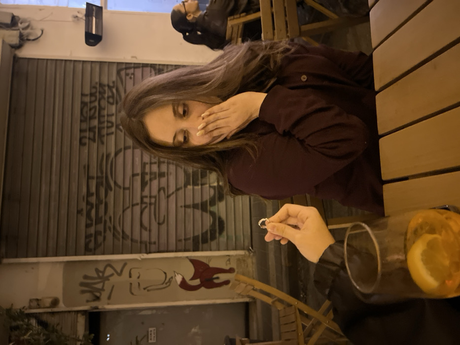 A woman seated at a wooden table reacts with surprise, covering her mouth with one hand, as a hand presents a ring. A glass of a yellow beverage sits nearby. Graffiti adorns a metal shutter in the background. Another person is partially visible in the upper part of the image. The scene appears to be outdoors at night.