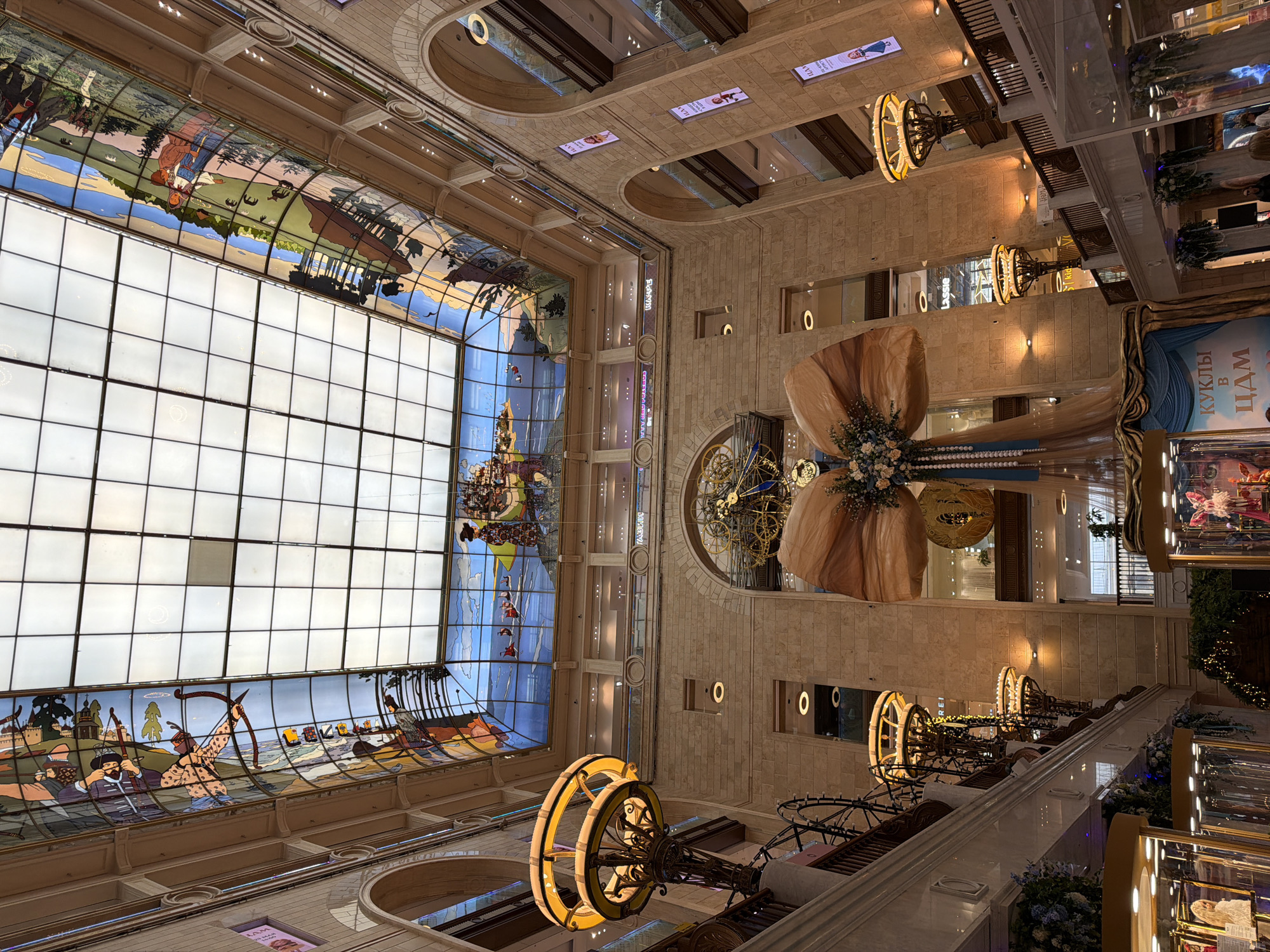 The image captures the interior of GUM (ГУМ), a large department store in Moscow, Russia, located on Red Square. The viewpoint is looking upwards. The ceiling features a large, gridded skylight, partially decorated with stained glass panels depicting historical Russian scenes. Below the skylight, a central architectural element of the building is visible, with ornate details and hanging decorations. Decorative chandeliers are suspended from the ceiling. Below the chandeliers are displays. The image conveys a sense of grandeur.